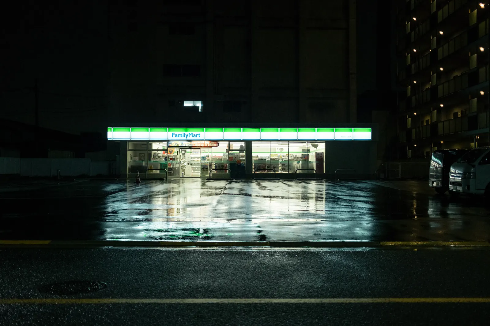 A FamilyMart glowing in the dark, its light reflected on the wet parking lot.
