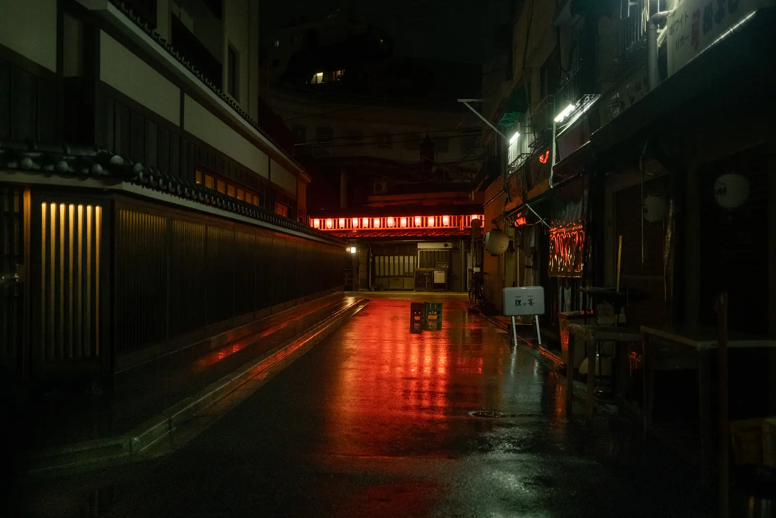 A narrow lane at night with red lanterns and wet pavement reflecting red light.