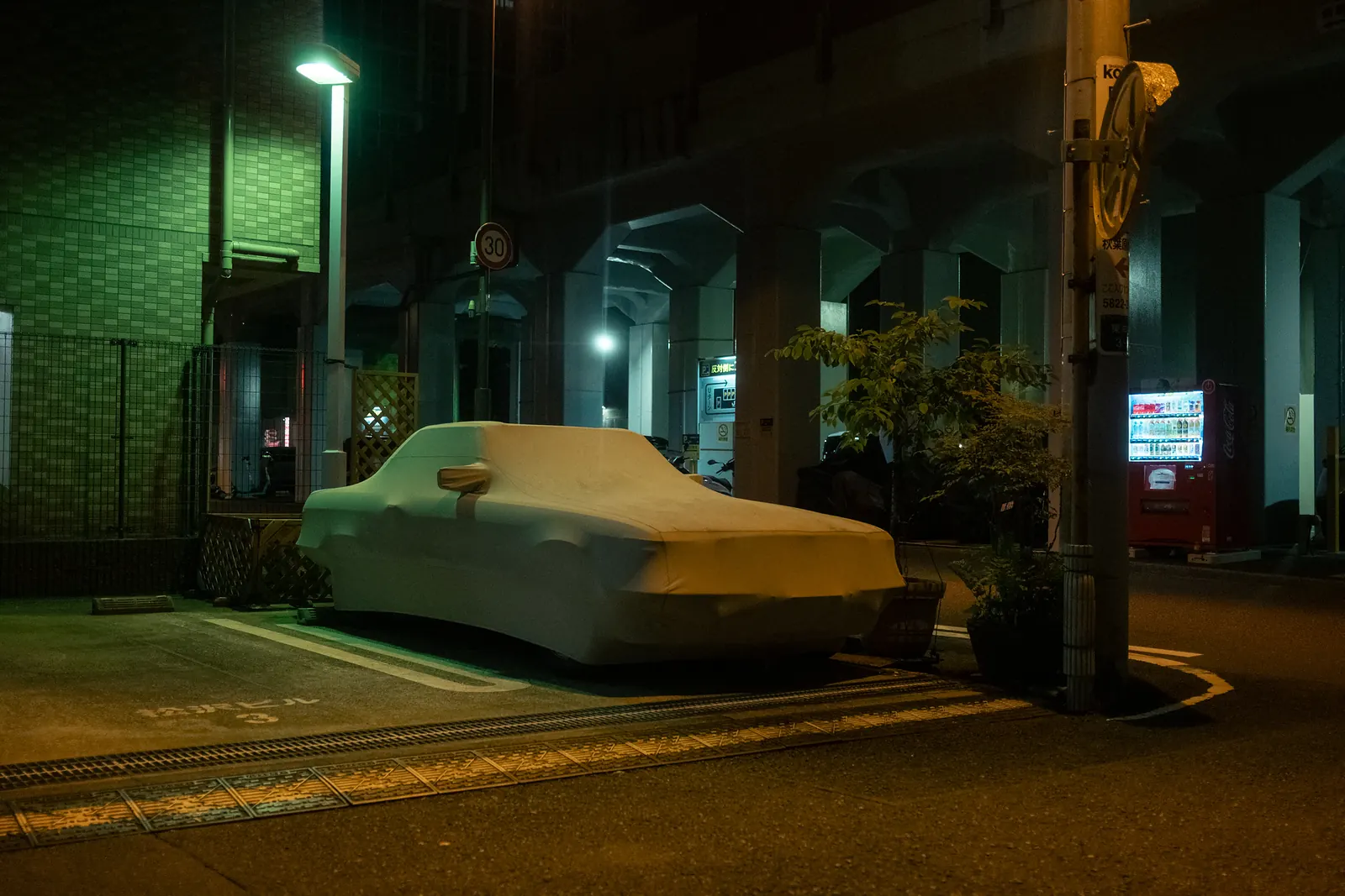 A covered car parked under an elevated highway at night, next to a vending machine.