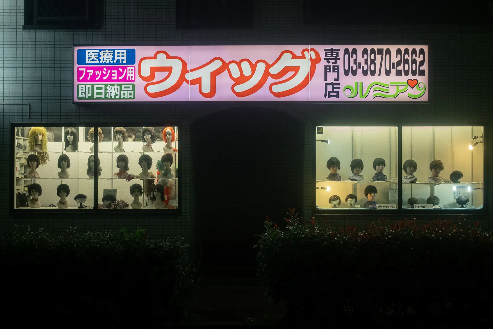A wig shop with lit windows and a neon sign on a dark facade.