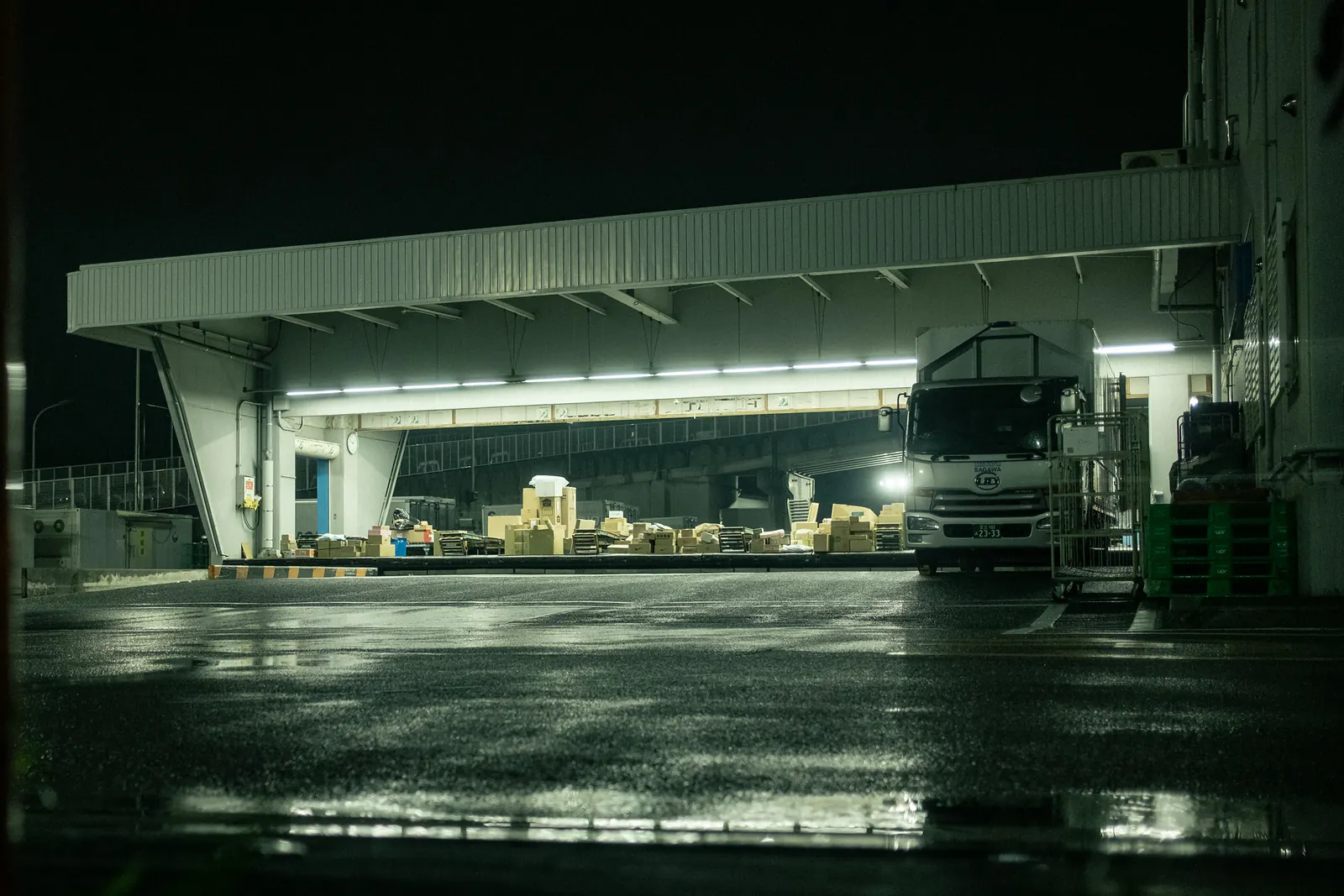 A loading dock lit by fluorescent tubes with a truck and stacked boxes, on wet asphalt.