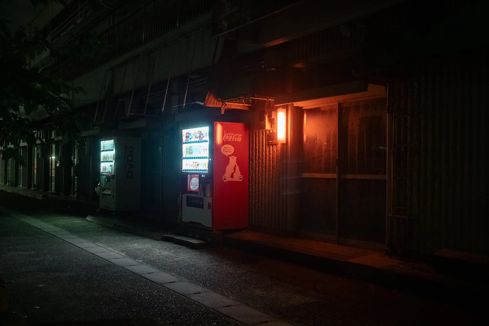 A Coca-Cola vending machine glowing beside a red-lit doorway on a dark lane.