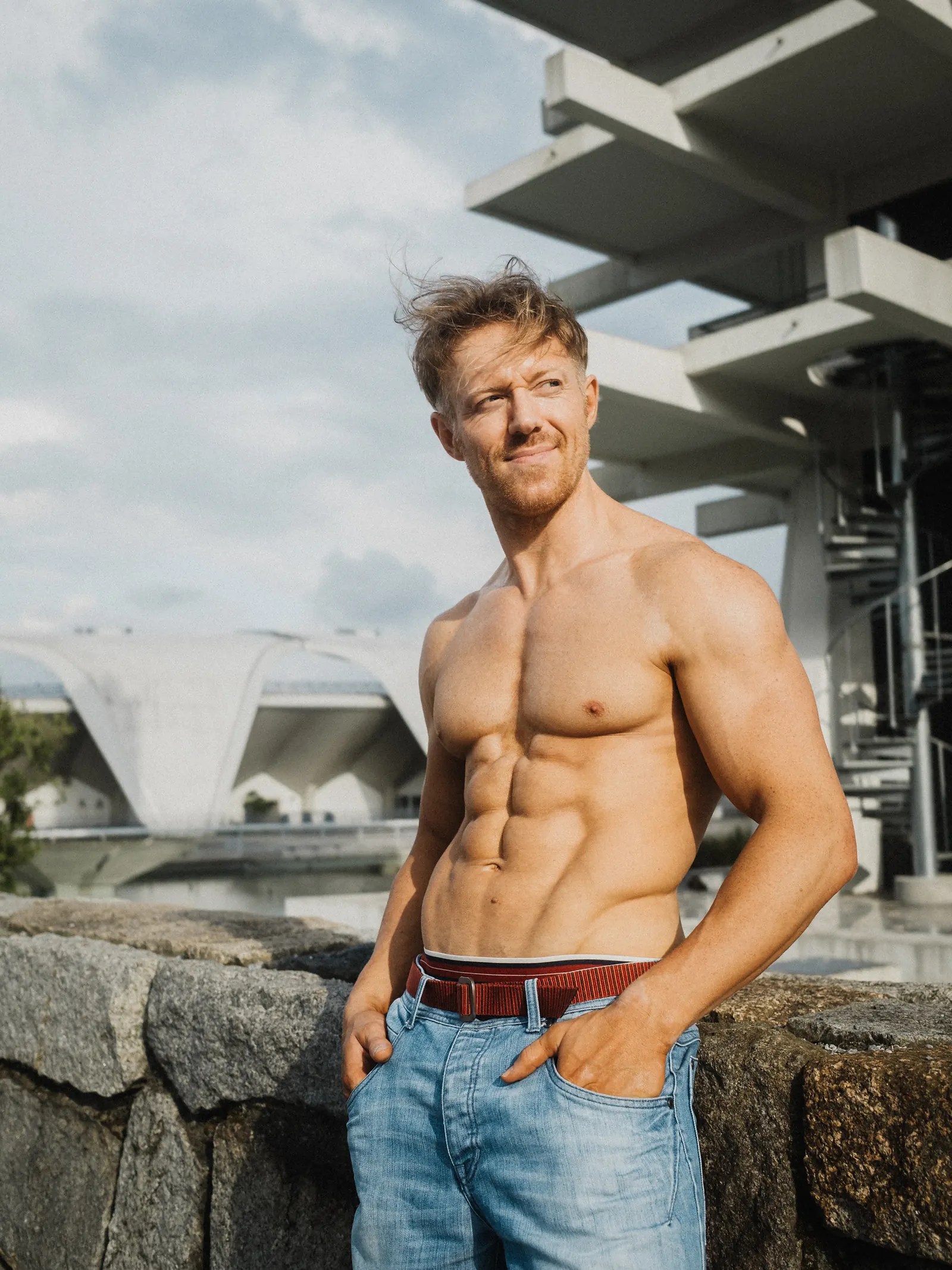 A shirtless man standing with his hands in his jeans pockets, looking off to the side, a concrete bridge and modern pavilion behind him.