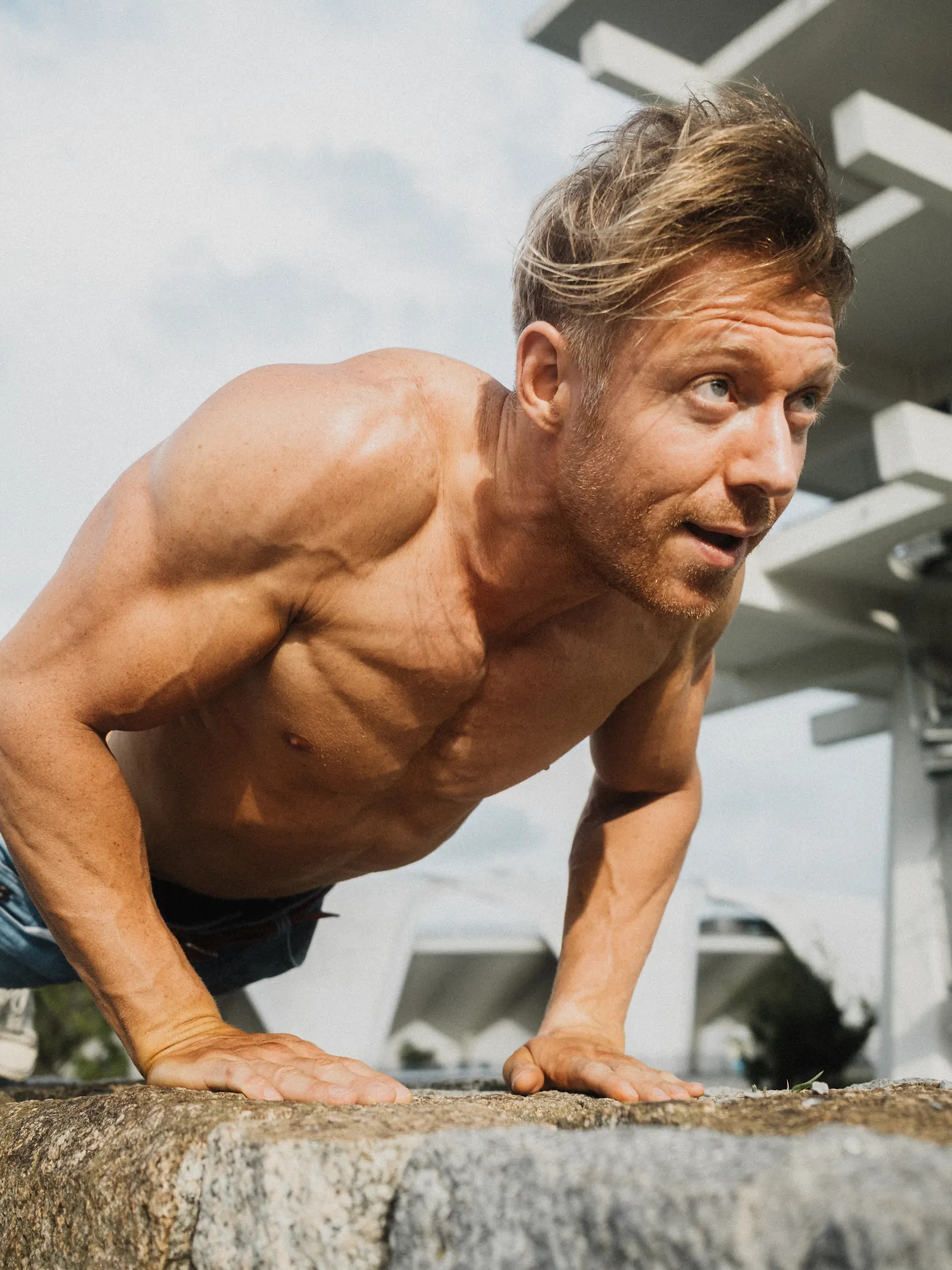 A man in a push-up position on a stone ledge, looking up and to the side, an angular concrete structure overhead.