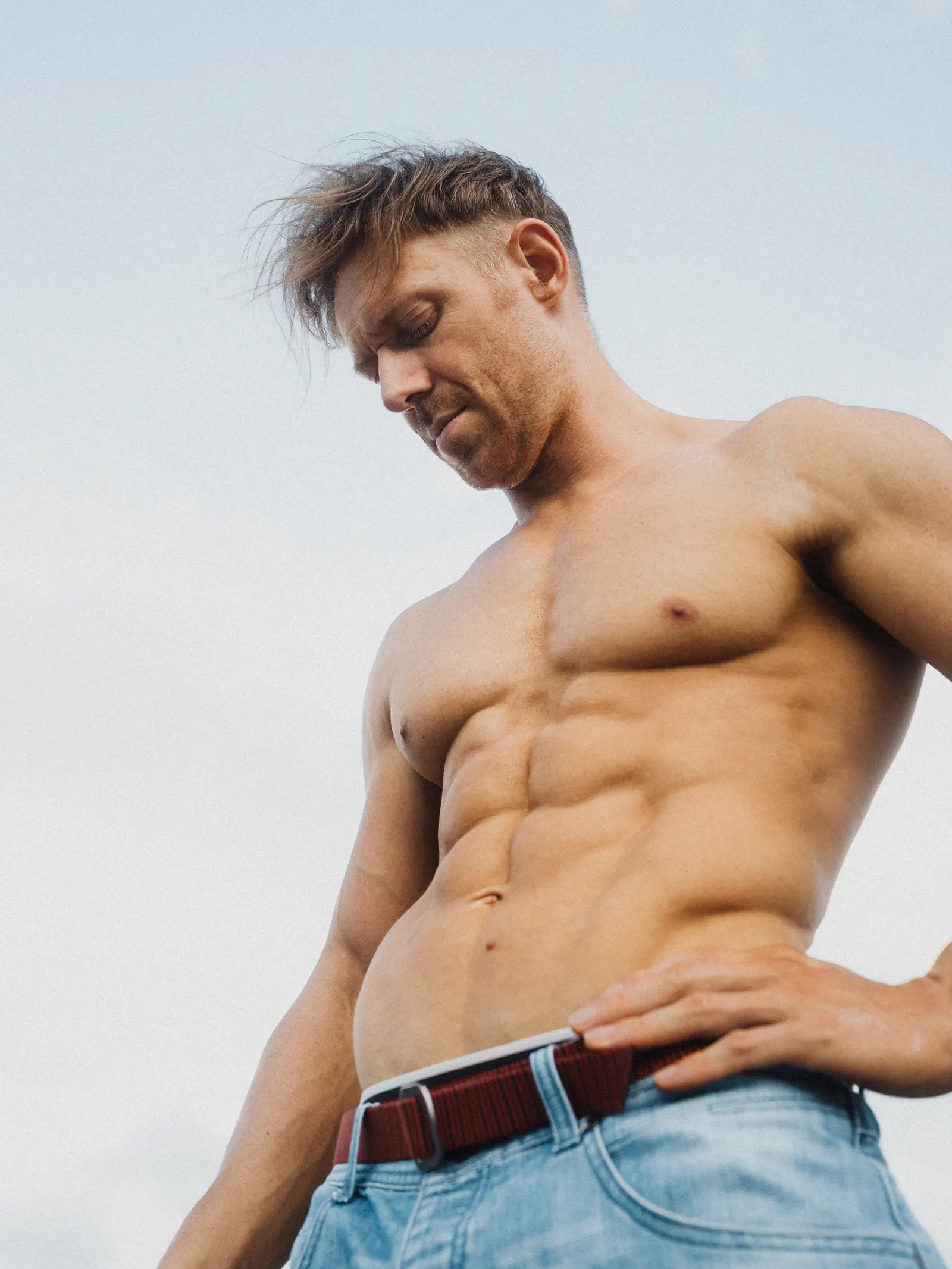 A man looking down at his torso, one hand resting on his belt, windswept hair against a plain sky.