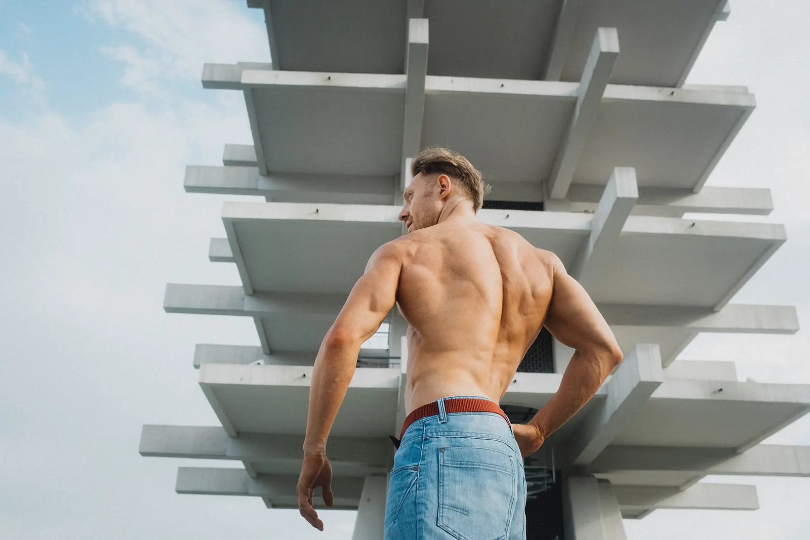 A man seen from behind, standing beneath a striking white geometric tower against the sky, one hand on his hip.