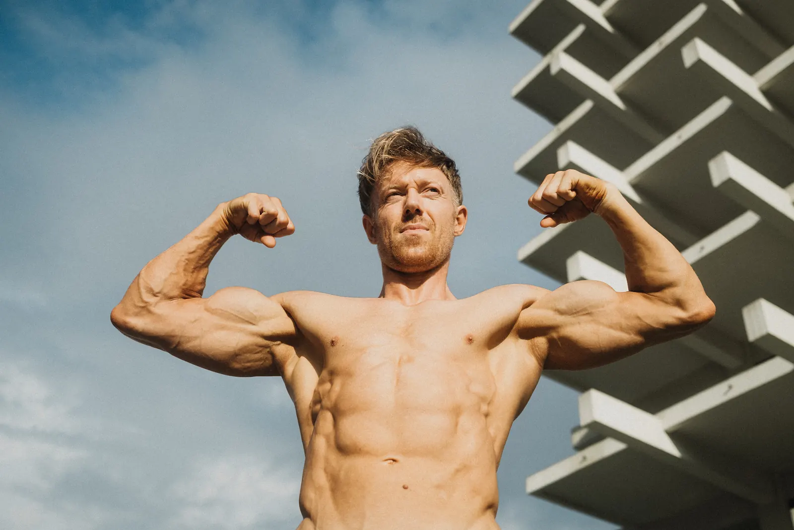A man flexing both biceps in a front double-bicep pose, looking upward, an angular white building behind him.
