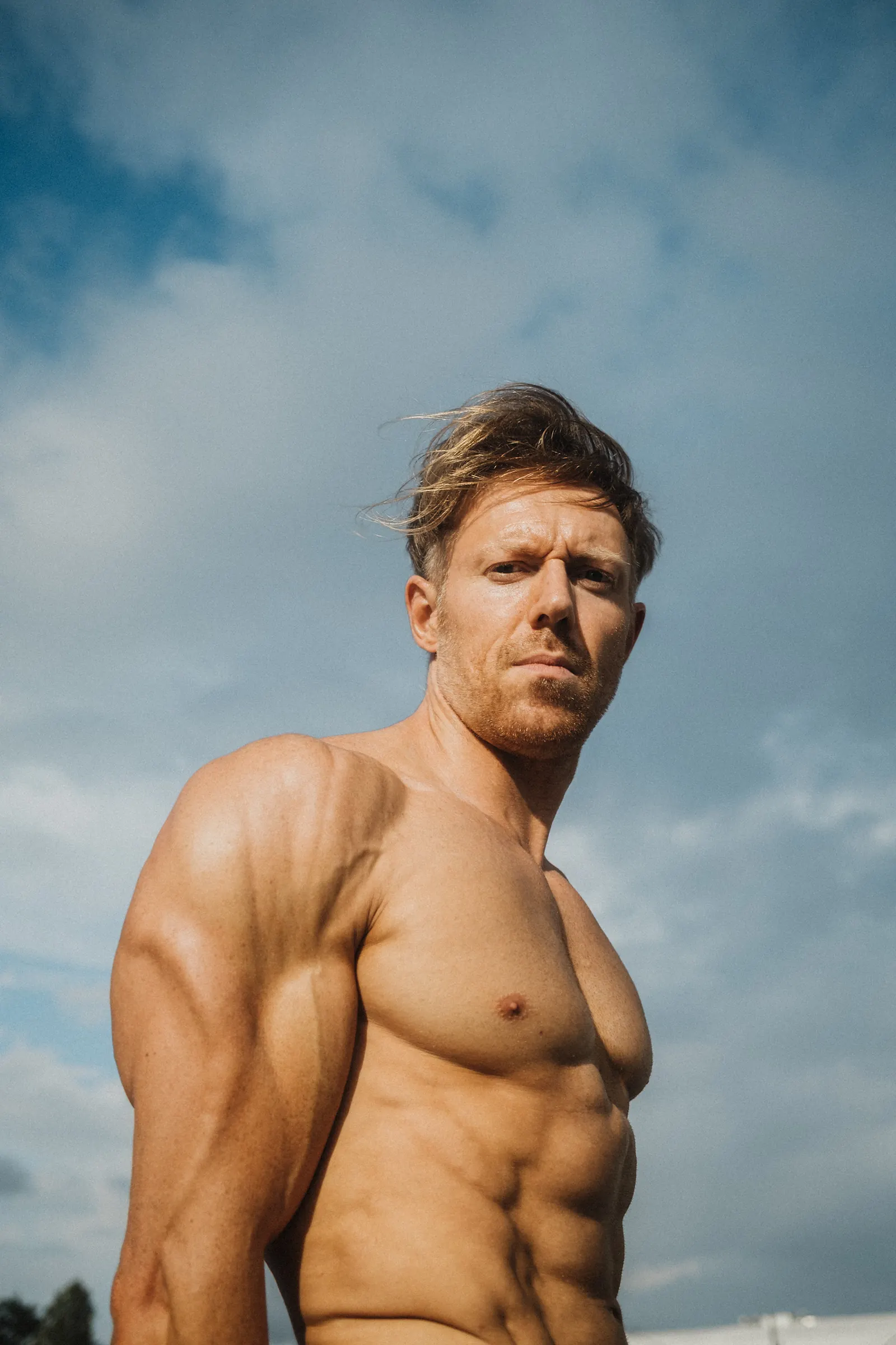 A man looking directly at the camera with a serious expression, sunlight on his shoulder against a cloudy sky.
