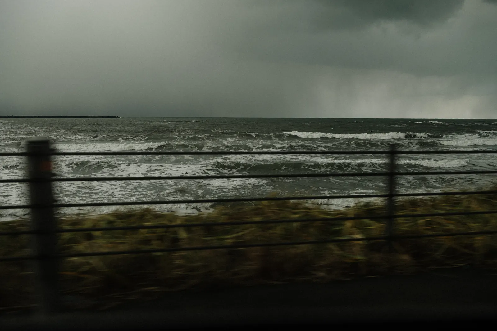 Waves and a fence blurred by speed, seen from a moving car on a stormy coast.