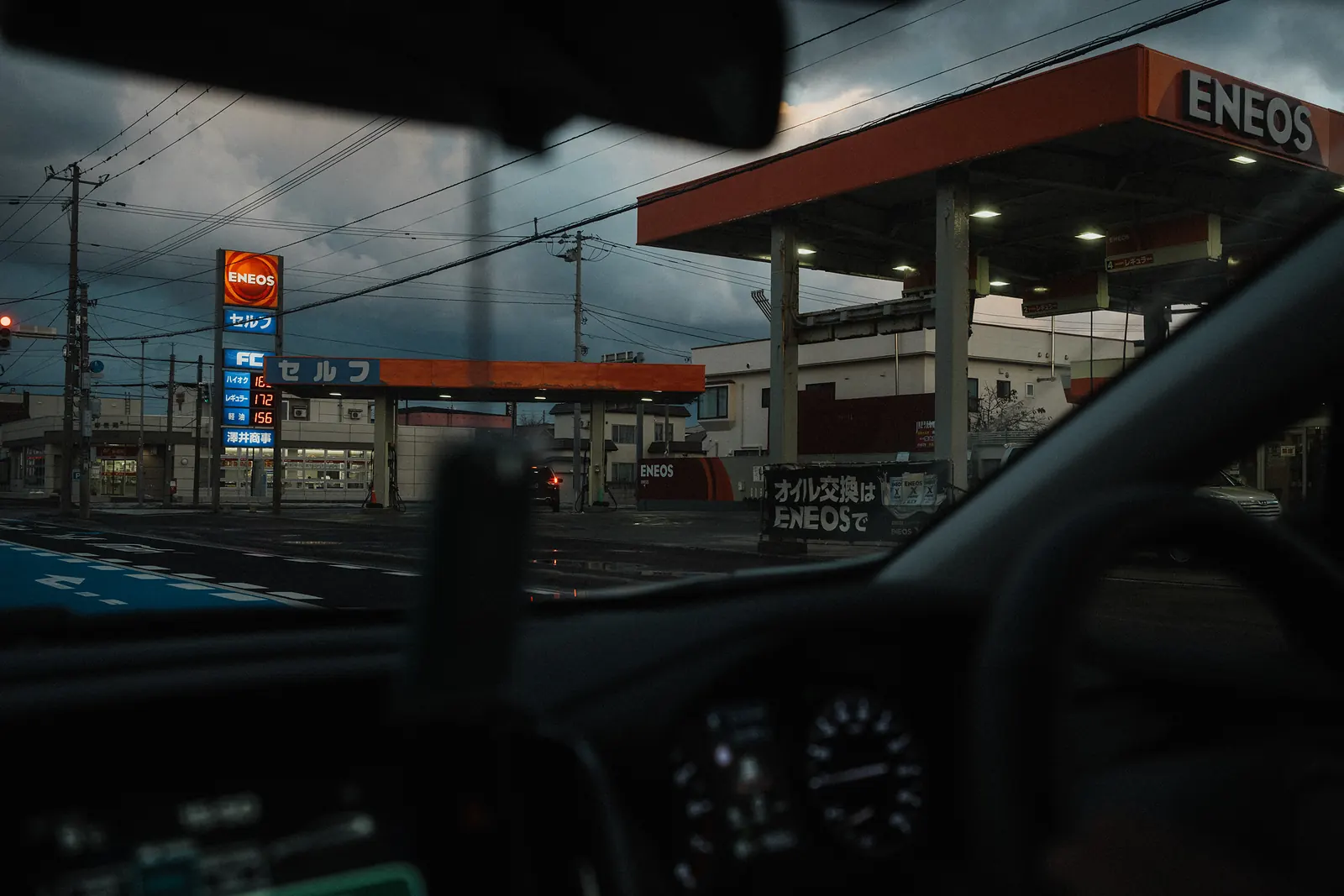 An ENEOS gas station glowing at dusk under dark clouds, seen through a car windshield.