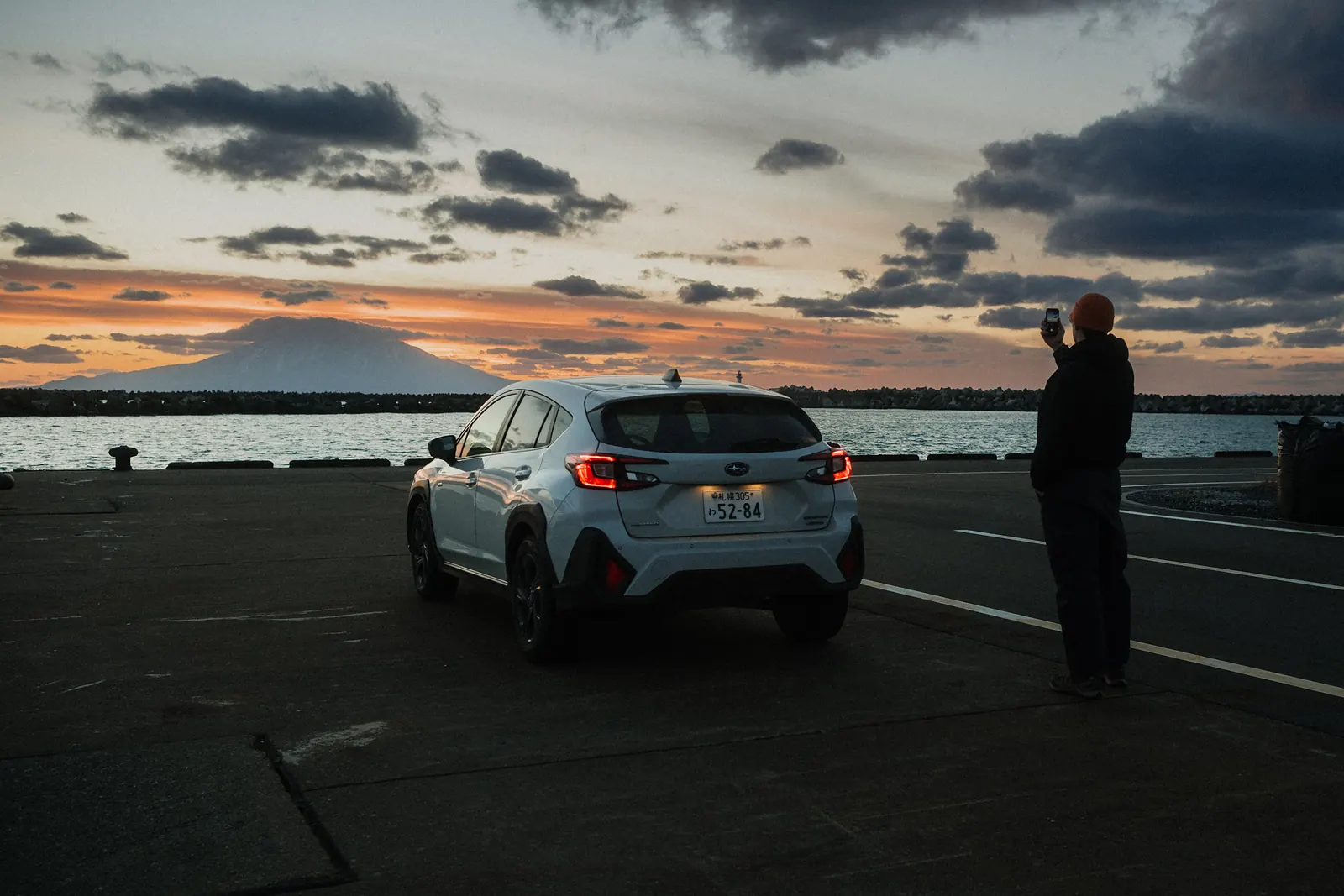 A white SUV and a man photographing the sunset behind a mountain across the harbor.