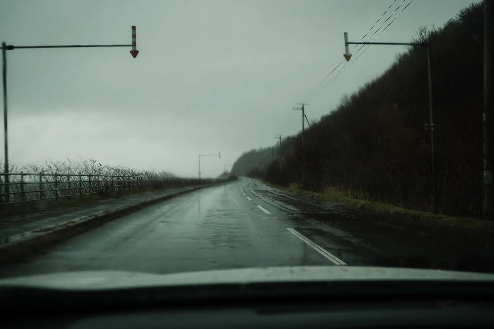 A wet road running past a dark hillside under a green-grey sky, seen through the windshield.