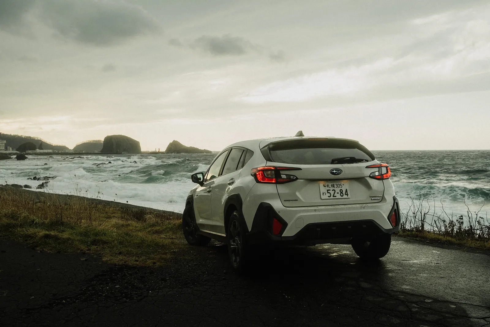 A white SUV parked beside a rocky, wave-battered coast in stormy light.