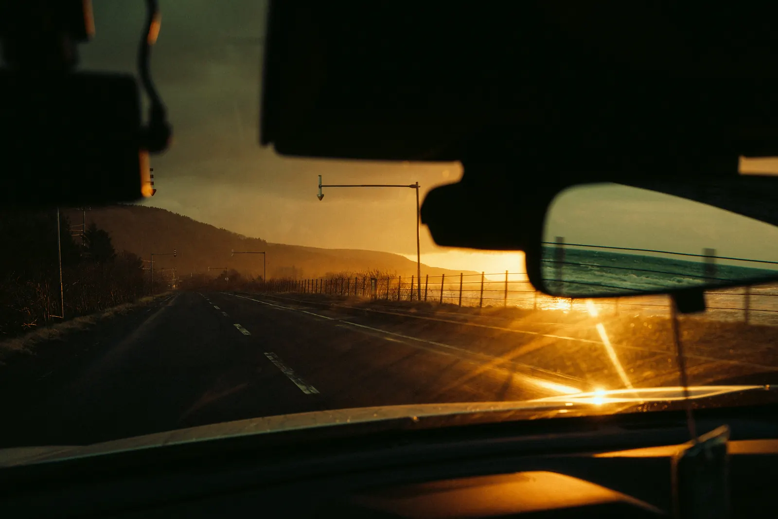 Low sun flaring across a coastal road, seen through the windshield.