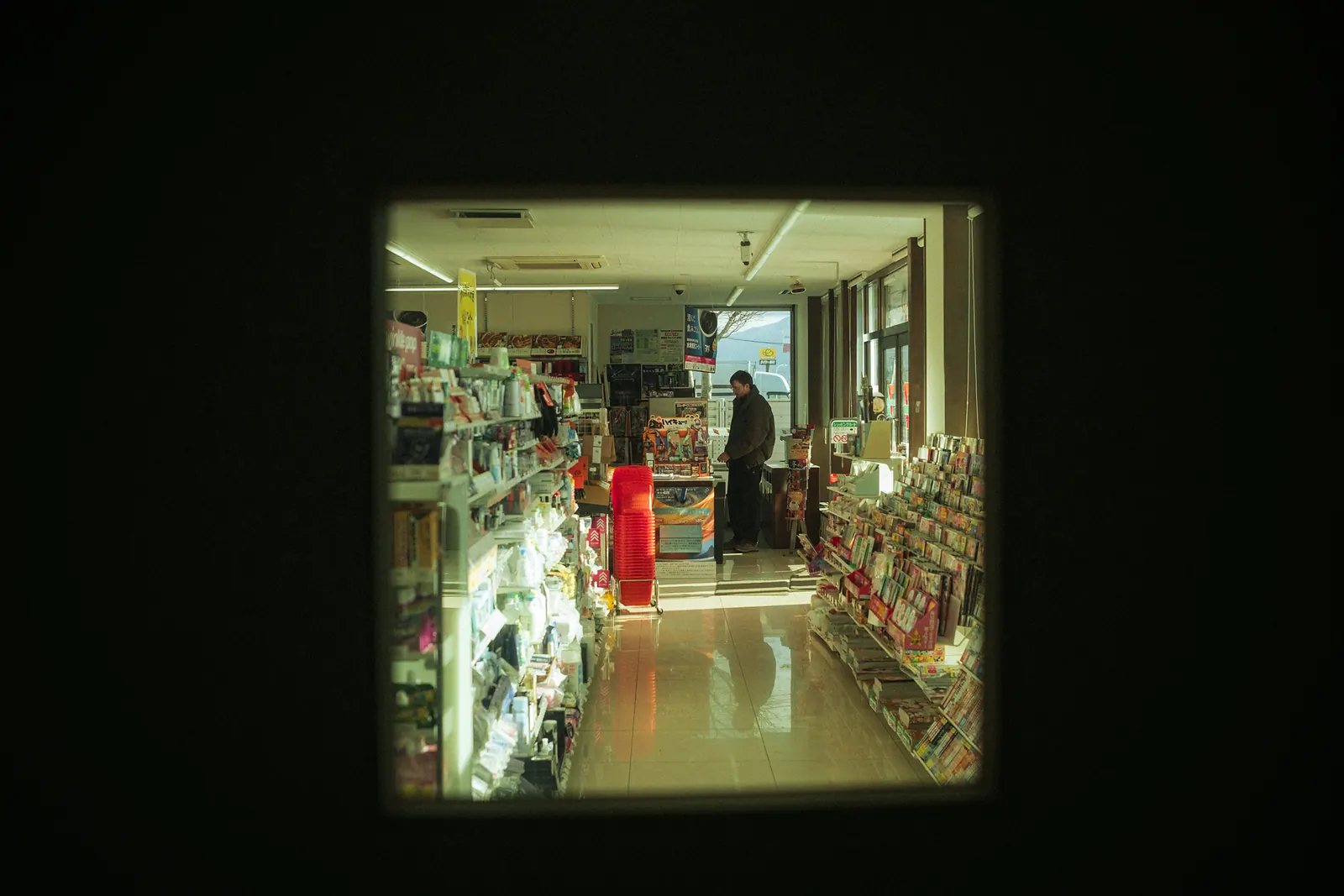 A convenience store interior glimpsed through a small opening in a dark wall.
