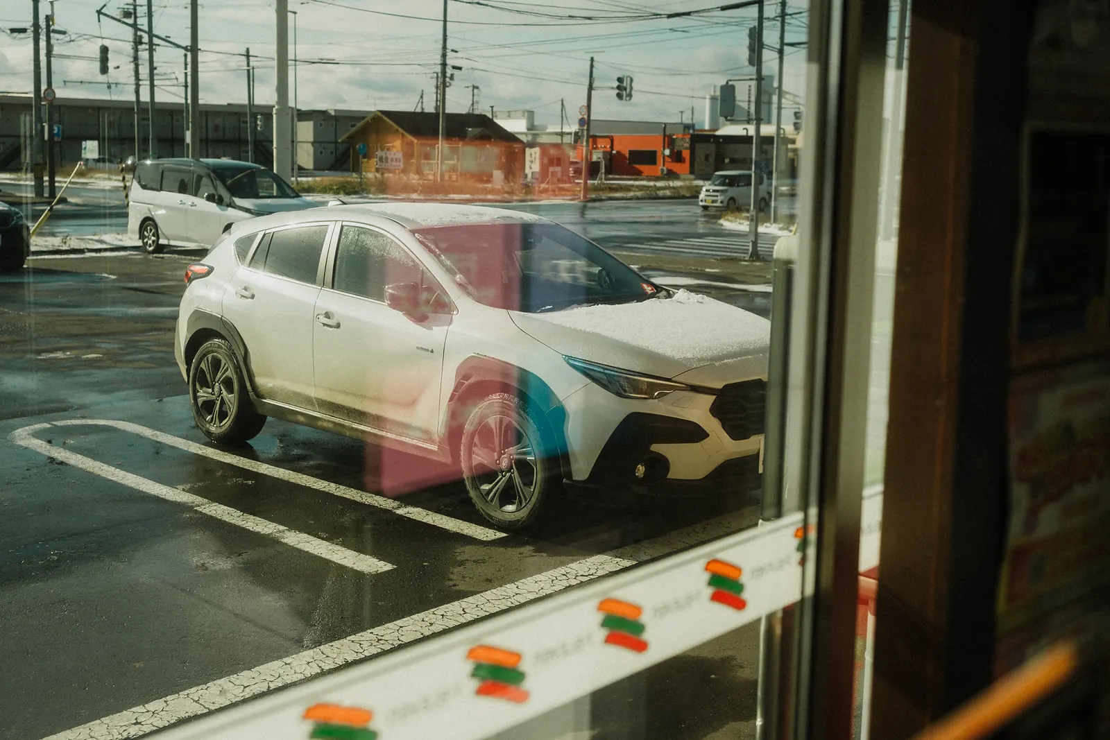A white car reflected in a convenience store window across a wet parking lot with patches of snow.