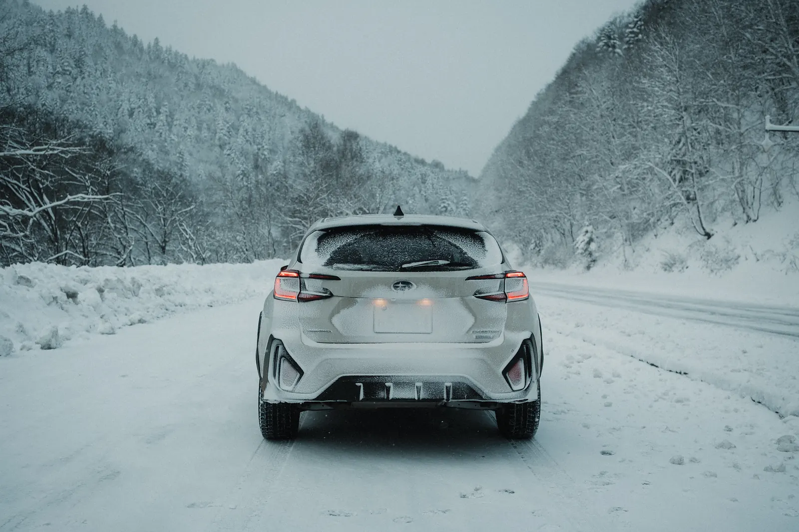 The rear of a snow-covered white SUV on a road between mountains.
