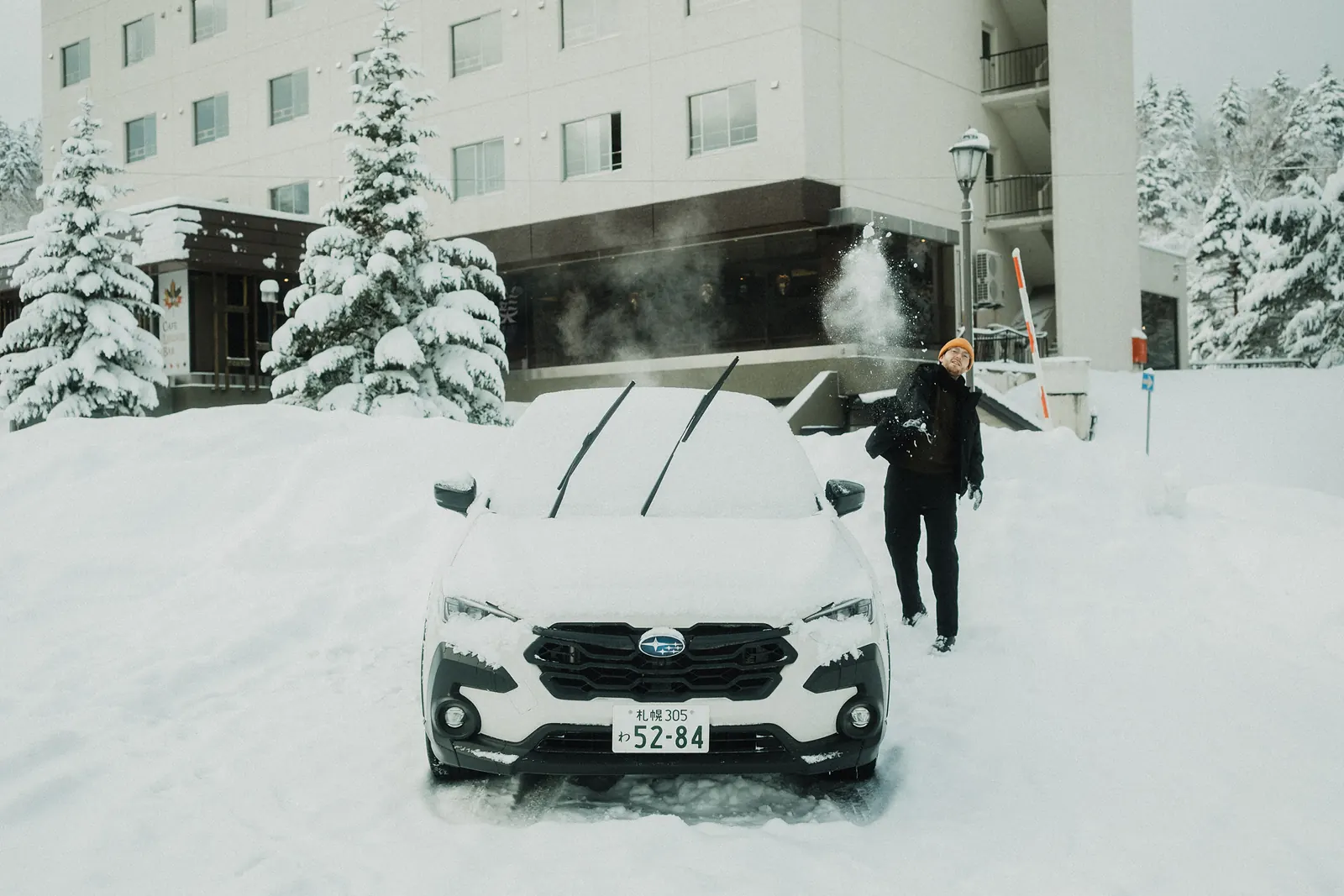 A man throwing snow beside a white SUV buried in snow outside a hotel.