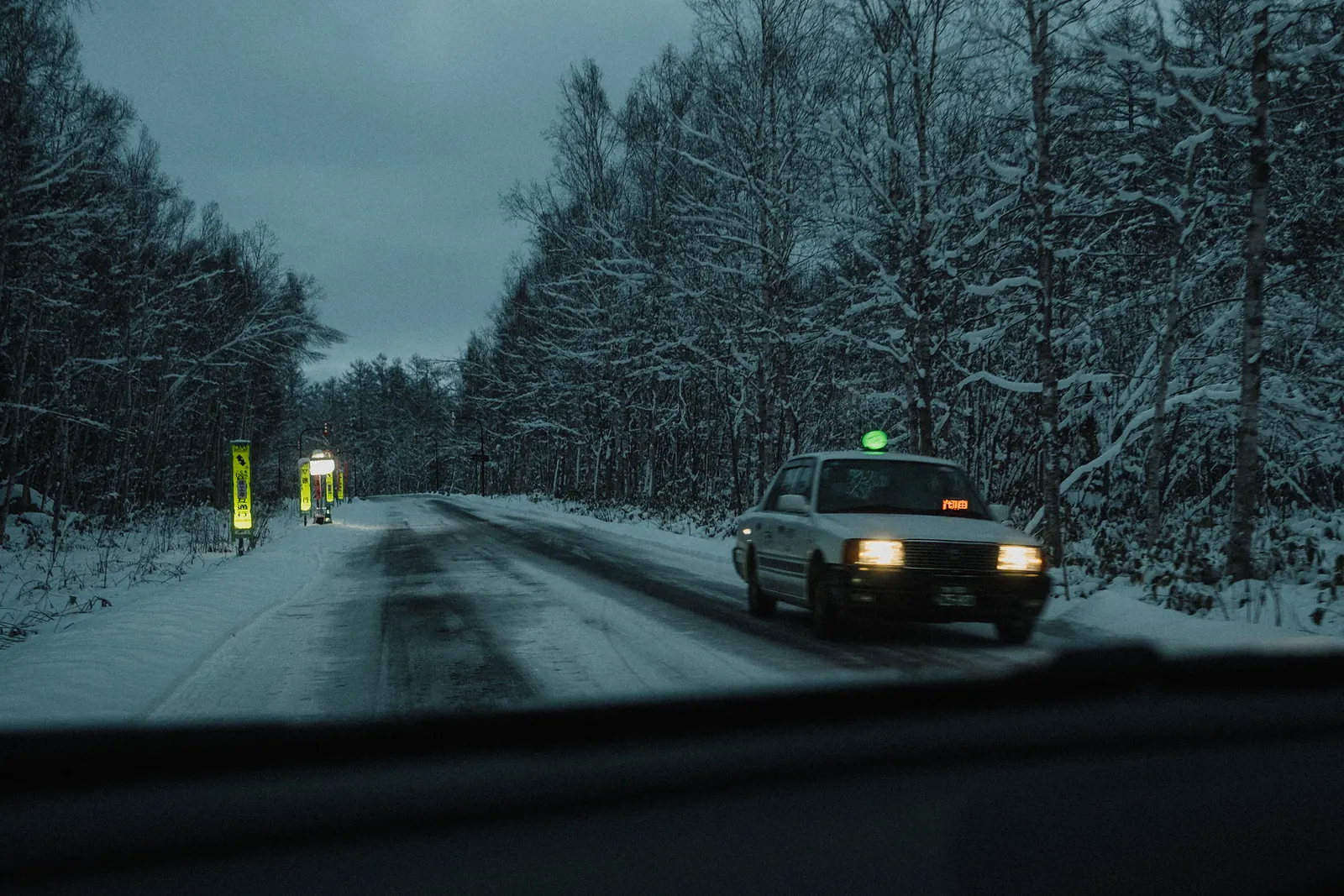 A taxi with its headlights on at dusk on a snowy forest road.