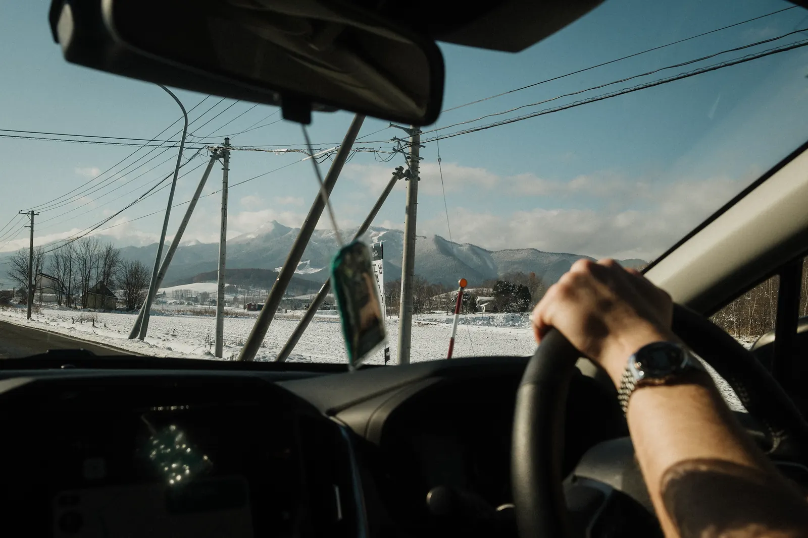 A hand with a wristwatch on the steering wheel, snowy mountains ahead.