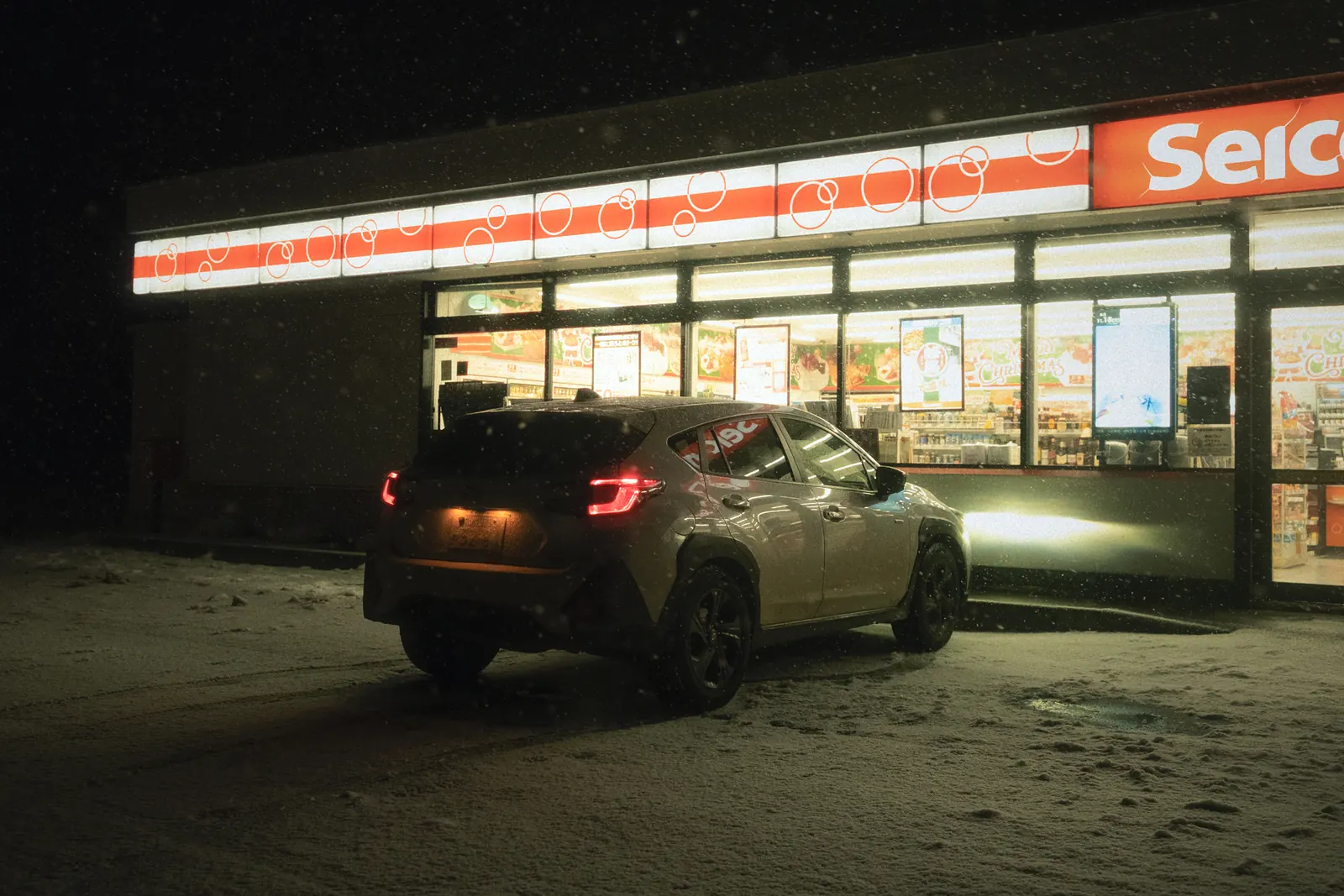 A car parked at night outside a glowing convenience store in falling snow.