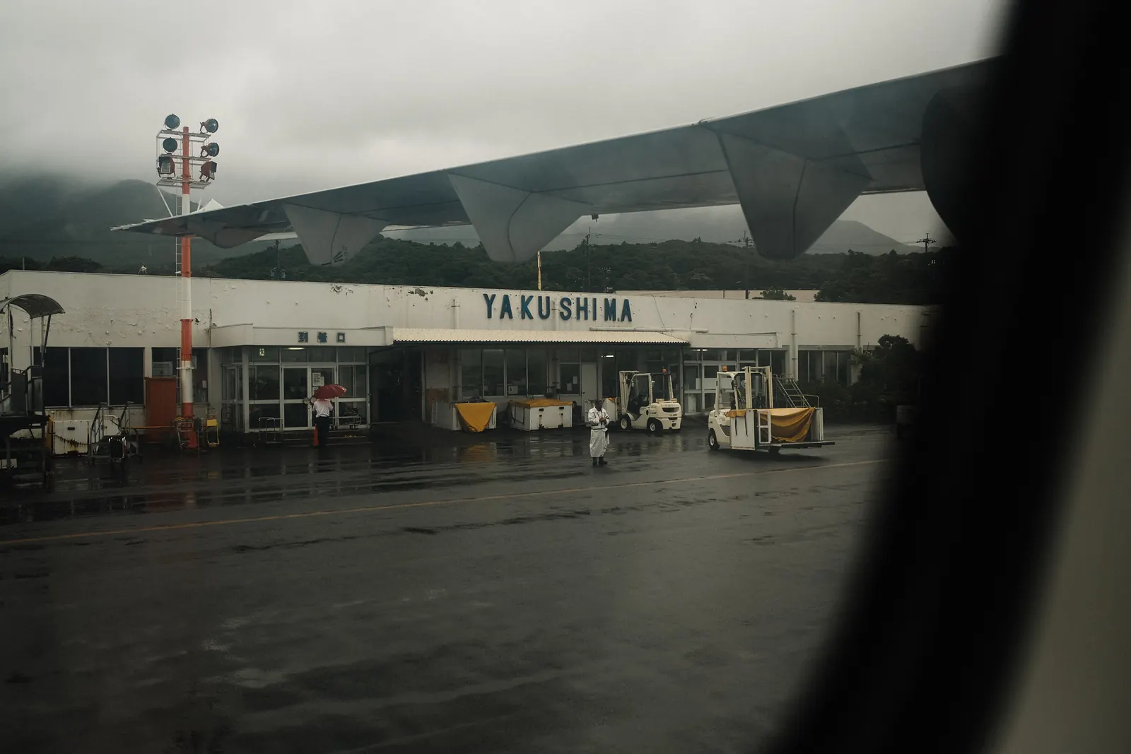 The Yakushima airport terminal under low cloud, wet tarmac in front of it, seen past the edge of a window.