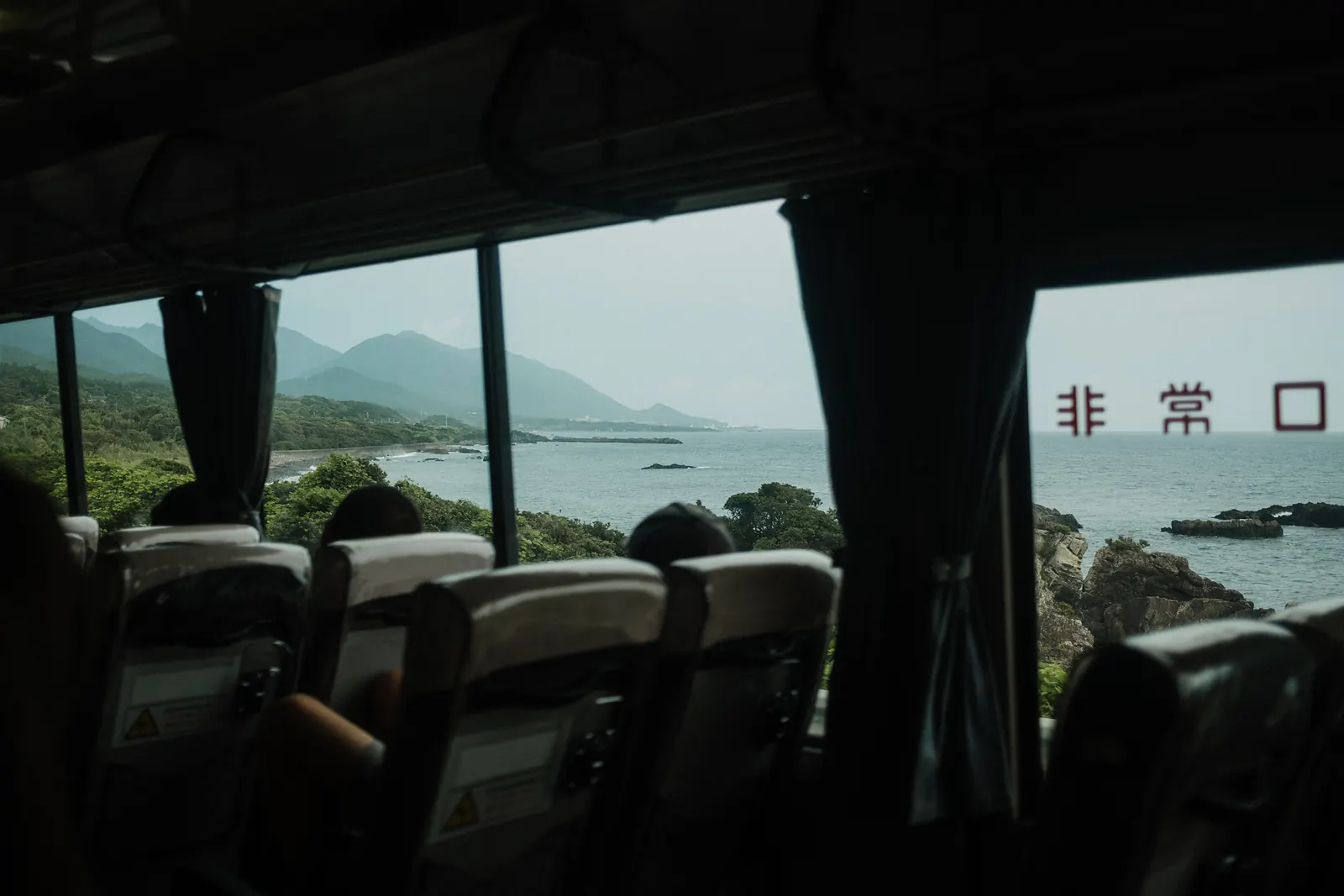 Seats inside a bus with the coast and distant mountains in the windows, an emergency-exit sign at the right.