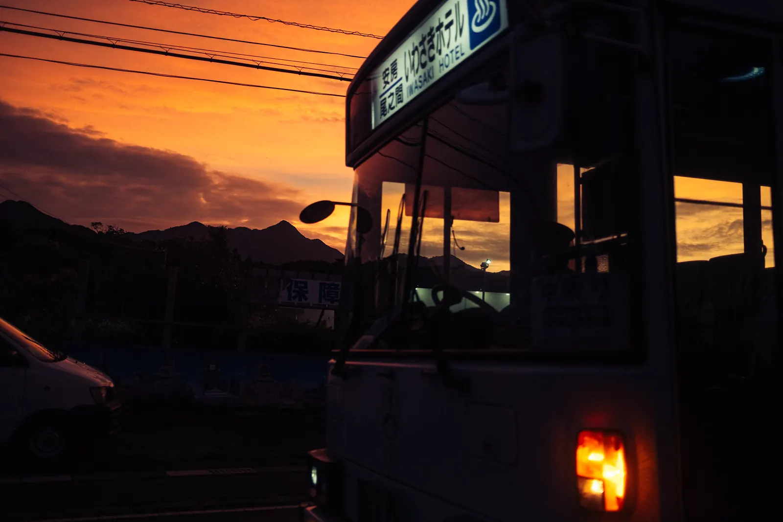 A bus pulling up in front of an orange sunset, its rear light glowing red, power lines overhead.