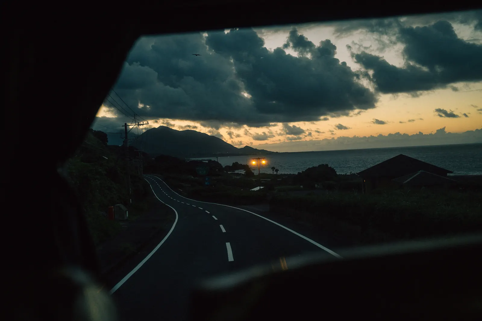 Dark clouds and a last strip of sunset over the sea, seen from a bus window above a curving coastal road.