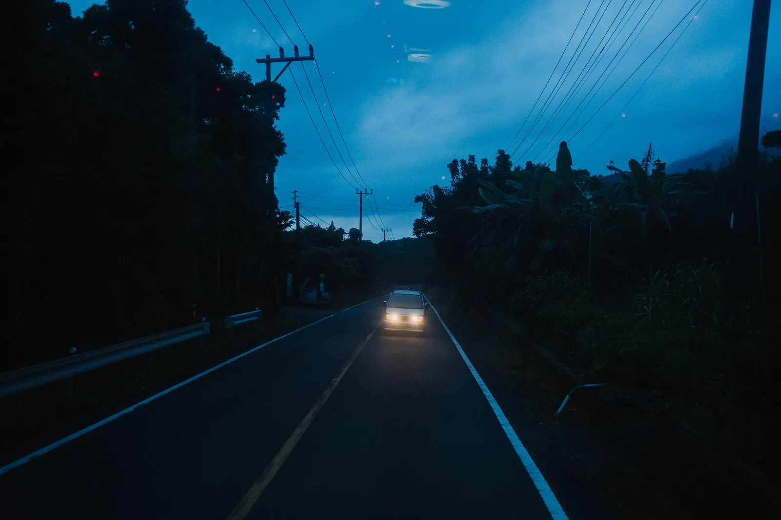 A single car with its headlights on coming along a coastal road under a blue dusk sky.