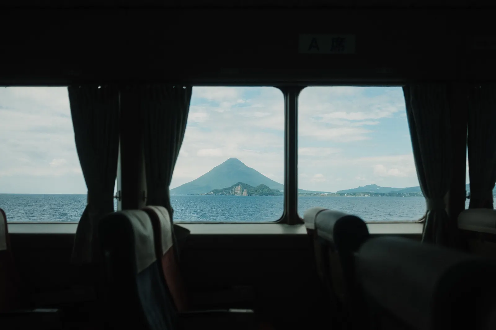 A conical mountain across the water, seen through the curtained windows of a bus.
