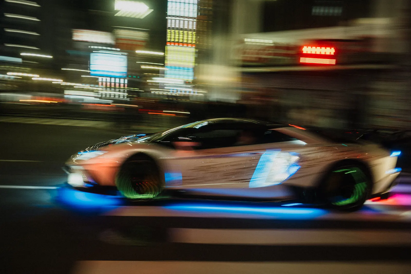 Motion-blurred Lamborghini wrapped in illuminated panels with glowing wheels and blue underglow, speeding past neon signs in Tokyo.