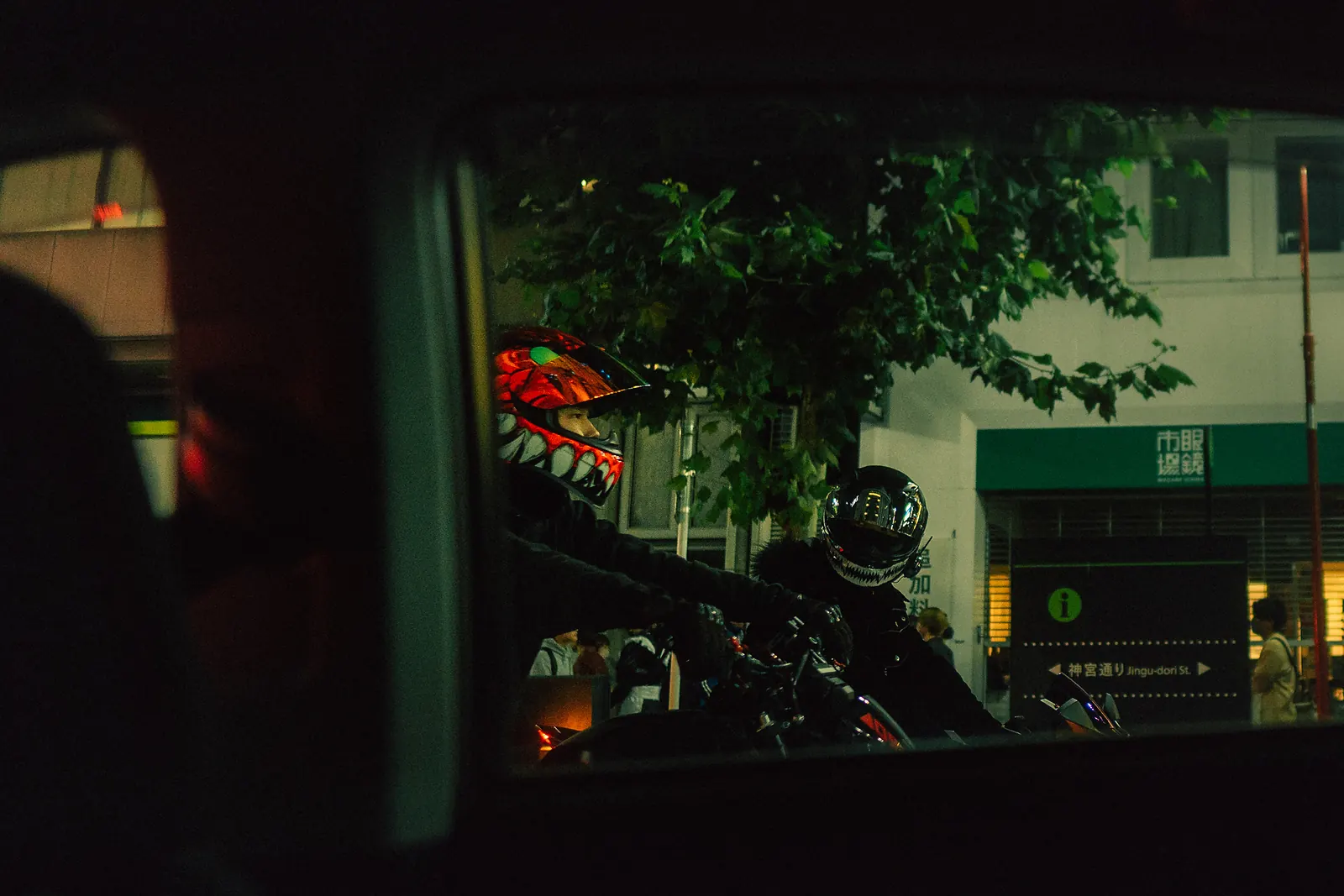 Motorcyclists at a Tokyo intersection seen in a car's side mirror, one wearing a red demon-faced helmet.