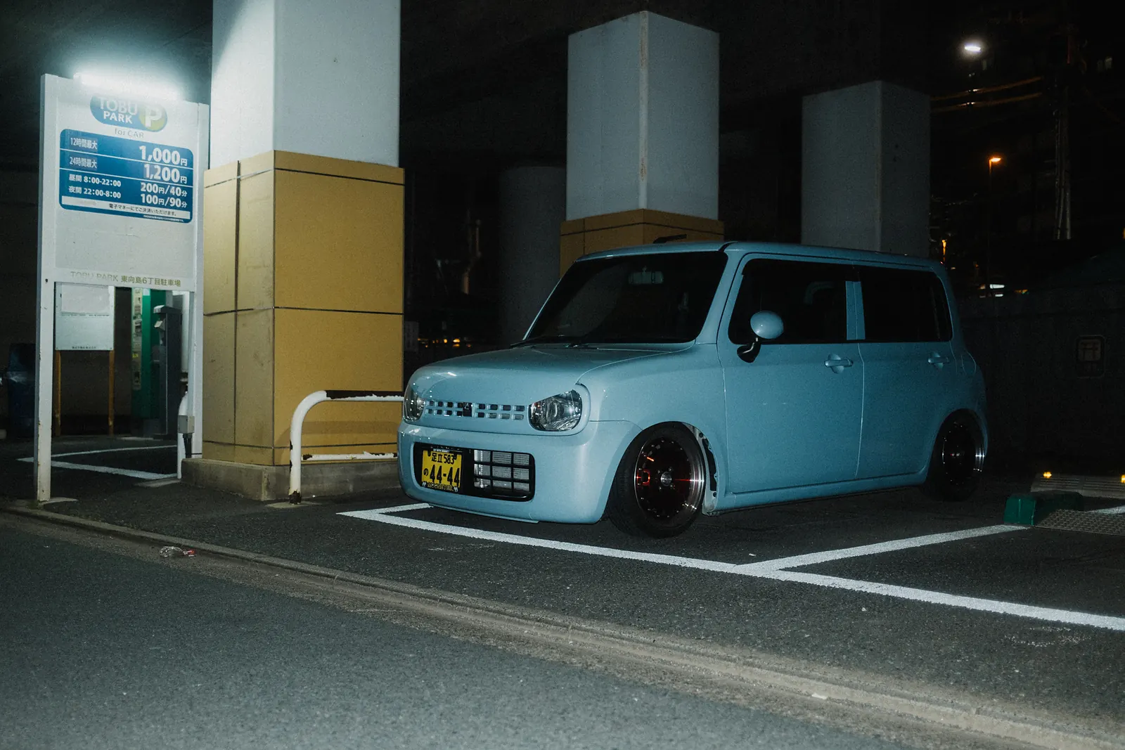 Pastel blue Suzuki Alto Lapin kei car on deep-dish wheels in a Tokyo coin parking lot at night.