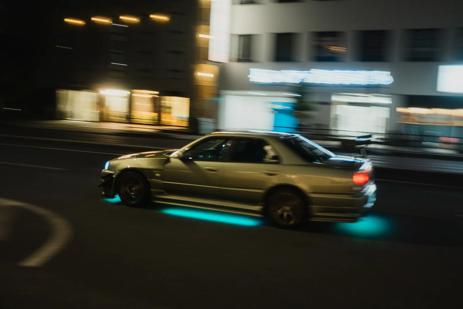 Silver Nissan Skyline sedan with turquoise underglow driving through Tokyo at night.