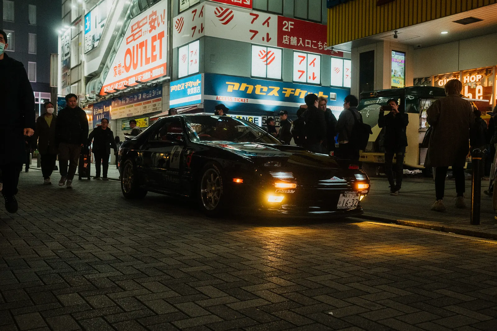 Black Mazda RX-7 with GReddy parts parked among pedestrians and shop signs in Akihabara, Tokyo.