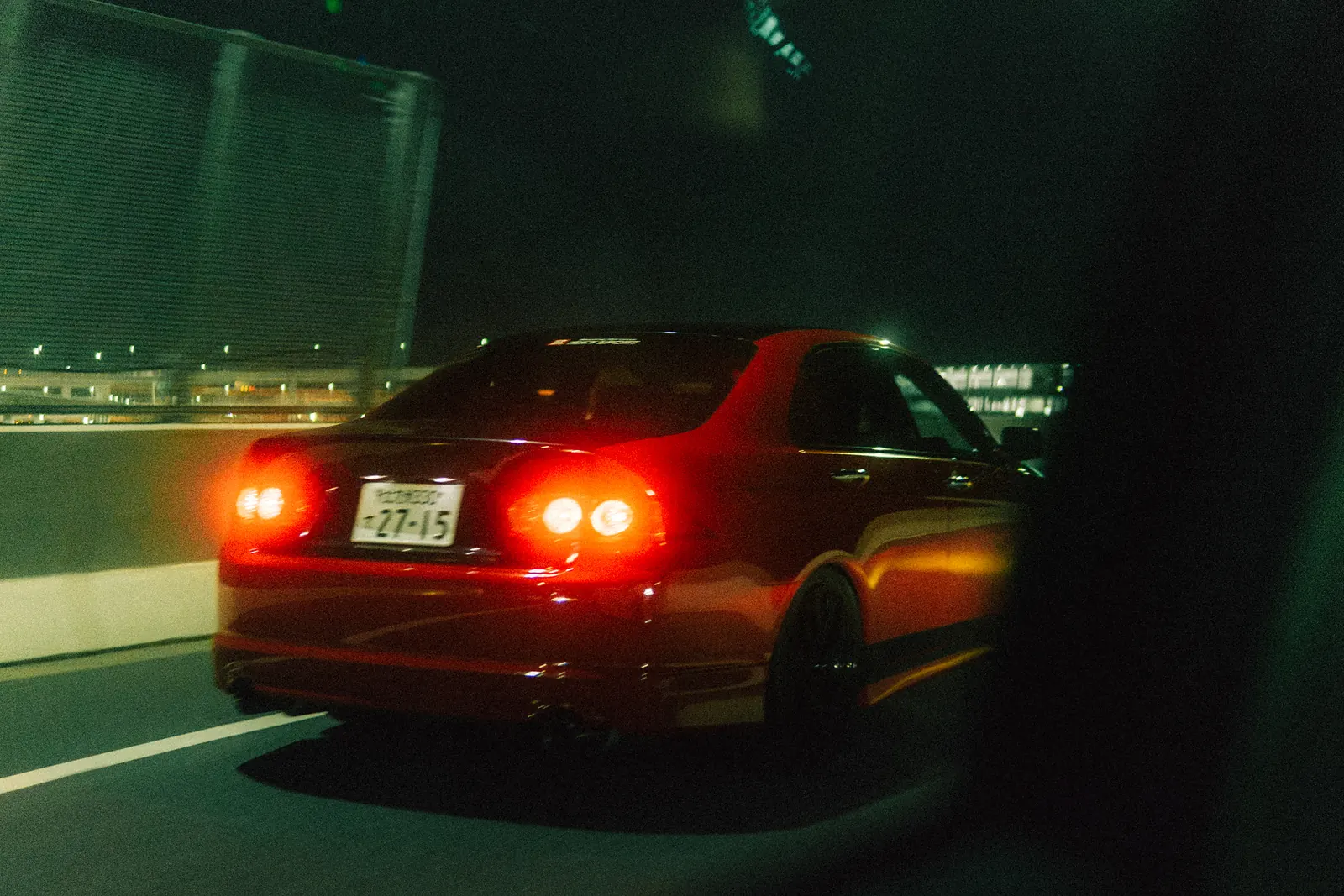 Red sedan with glowing taillights on a Tokyo expressway at night, shot from a moving car.
