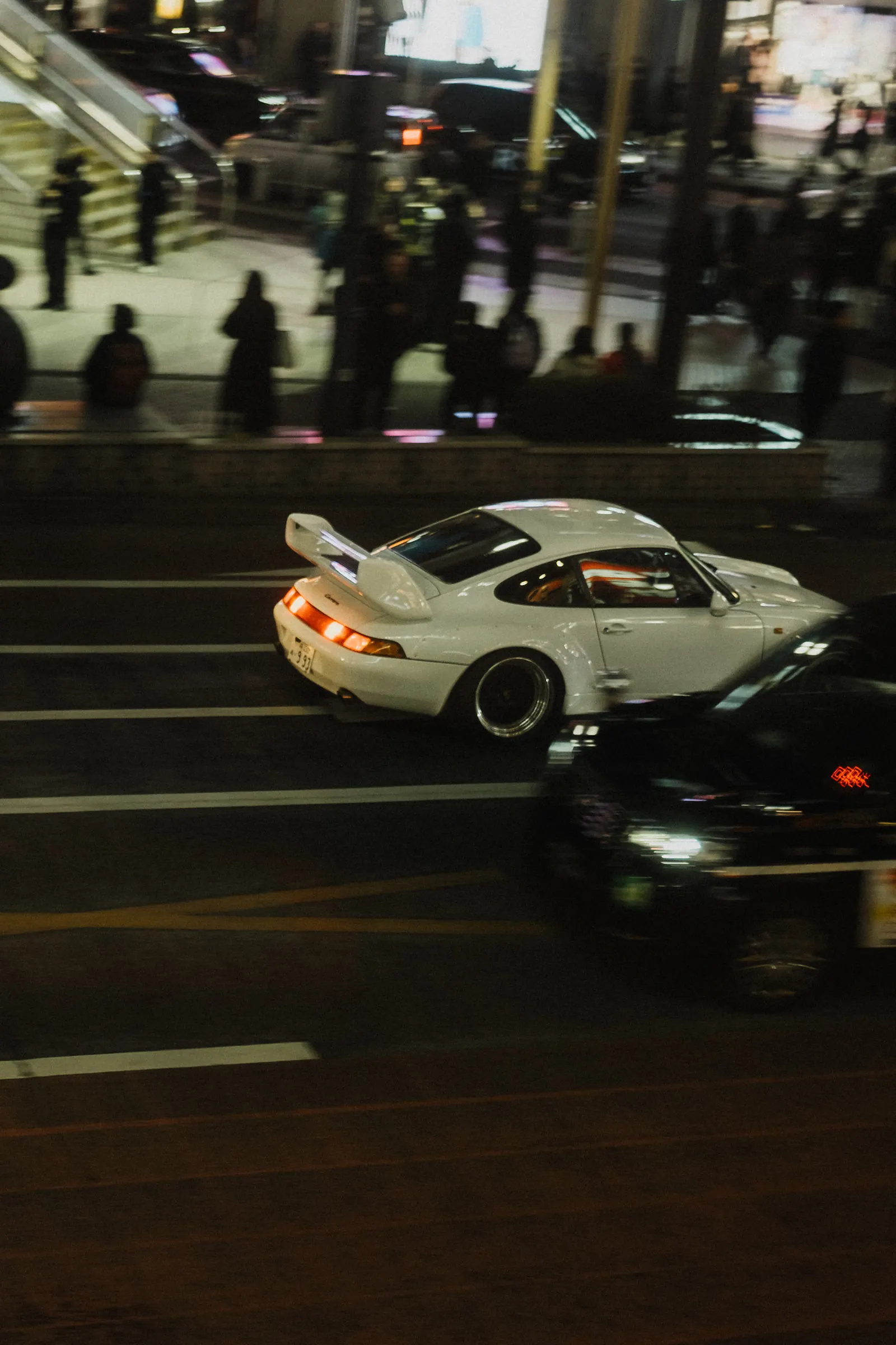 White RWB Porsche 911 widebody driving through Shibuya at night, seen from above with crowds in the background.