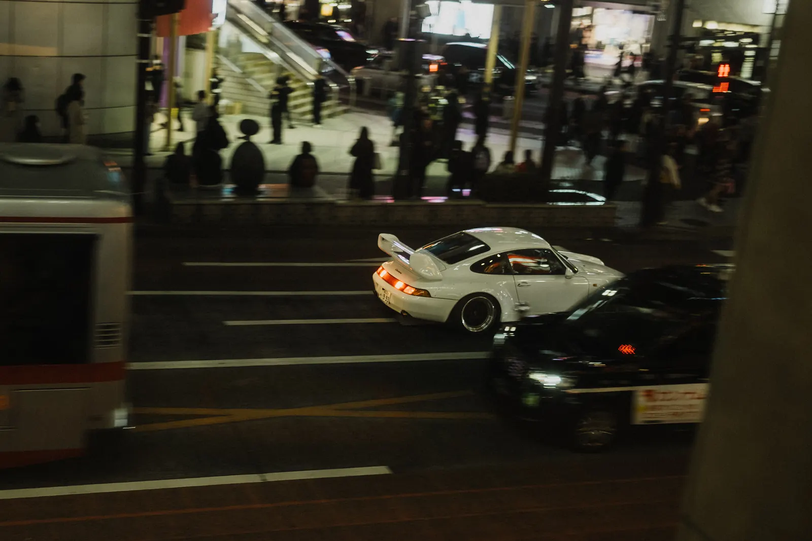 White RWB Porsche 911 widebody driving through Shibuya at night, seen from above with crowds in the background.