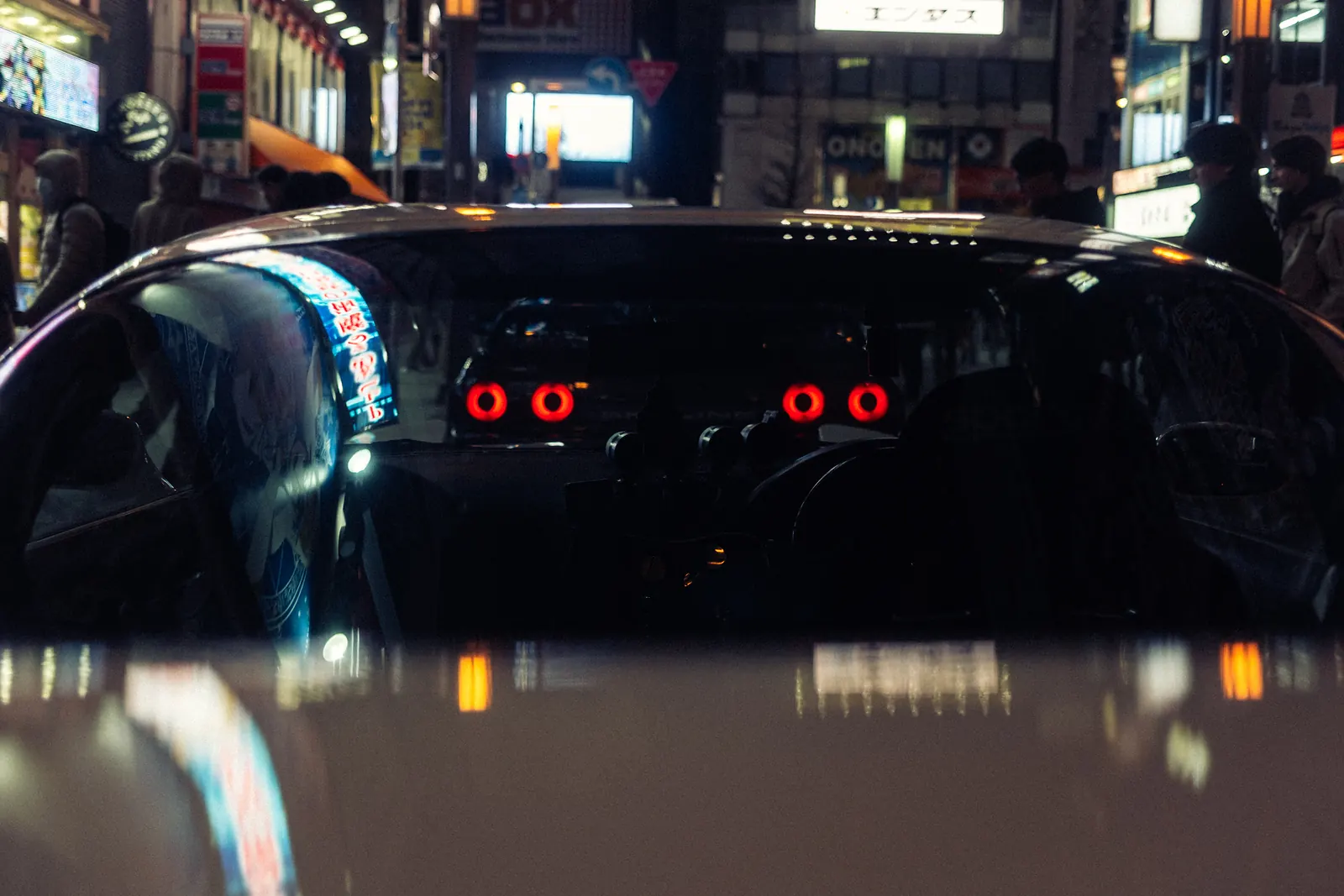 Neon reflections on a windshield framing the round taillights of a Nissan Skyline GT-R V-Spec II in Tokyo at night.