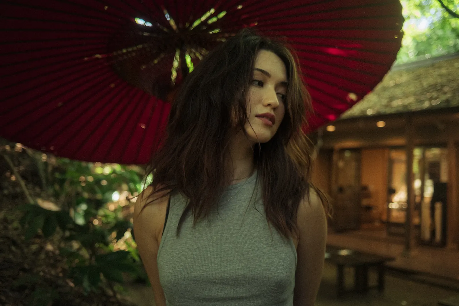 A woman with tousled dark hair looking down and away, framed by a large red paper parasol, a wooden teahouse blurred behind her.