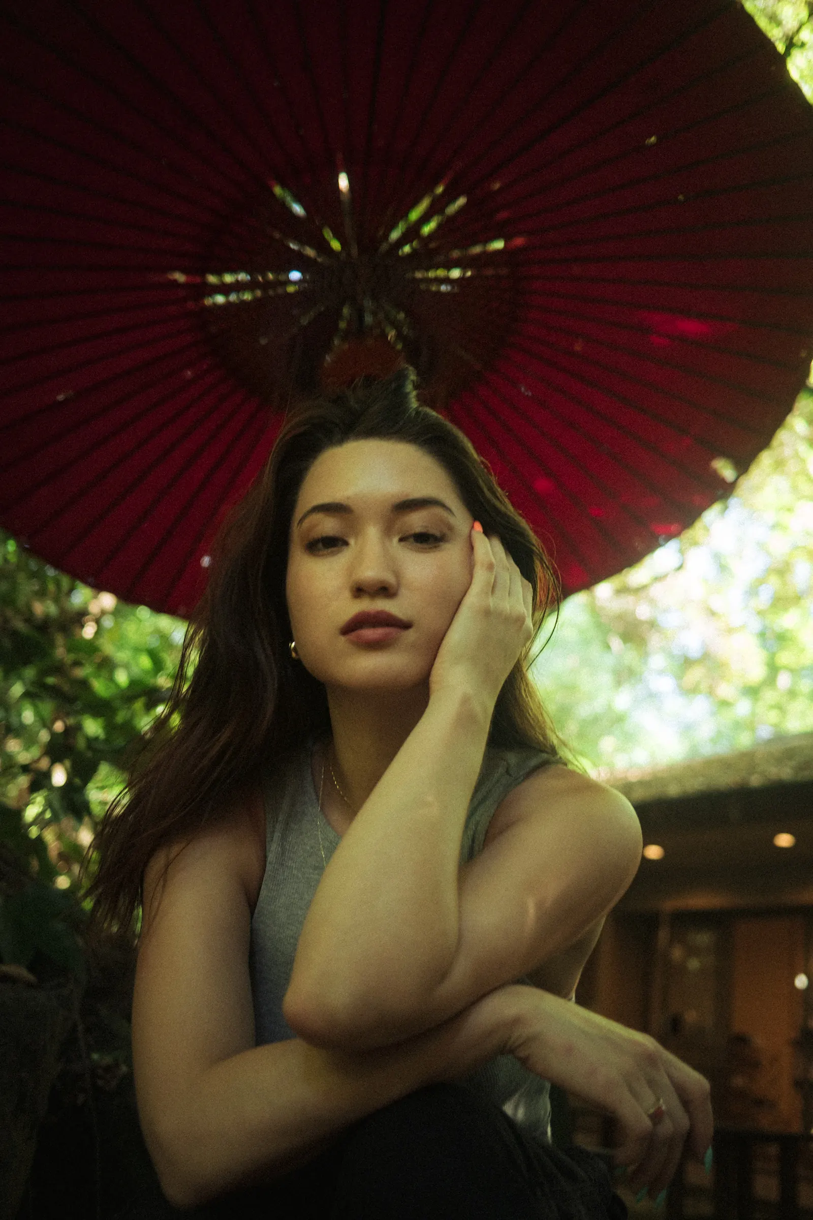A woman resting her cheek against her hand, looking into the camera from below a large red paper parasol, green foliage around her.