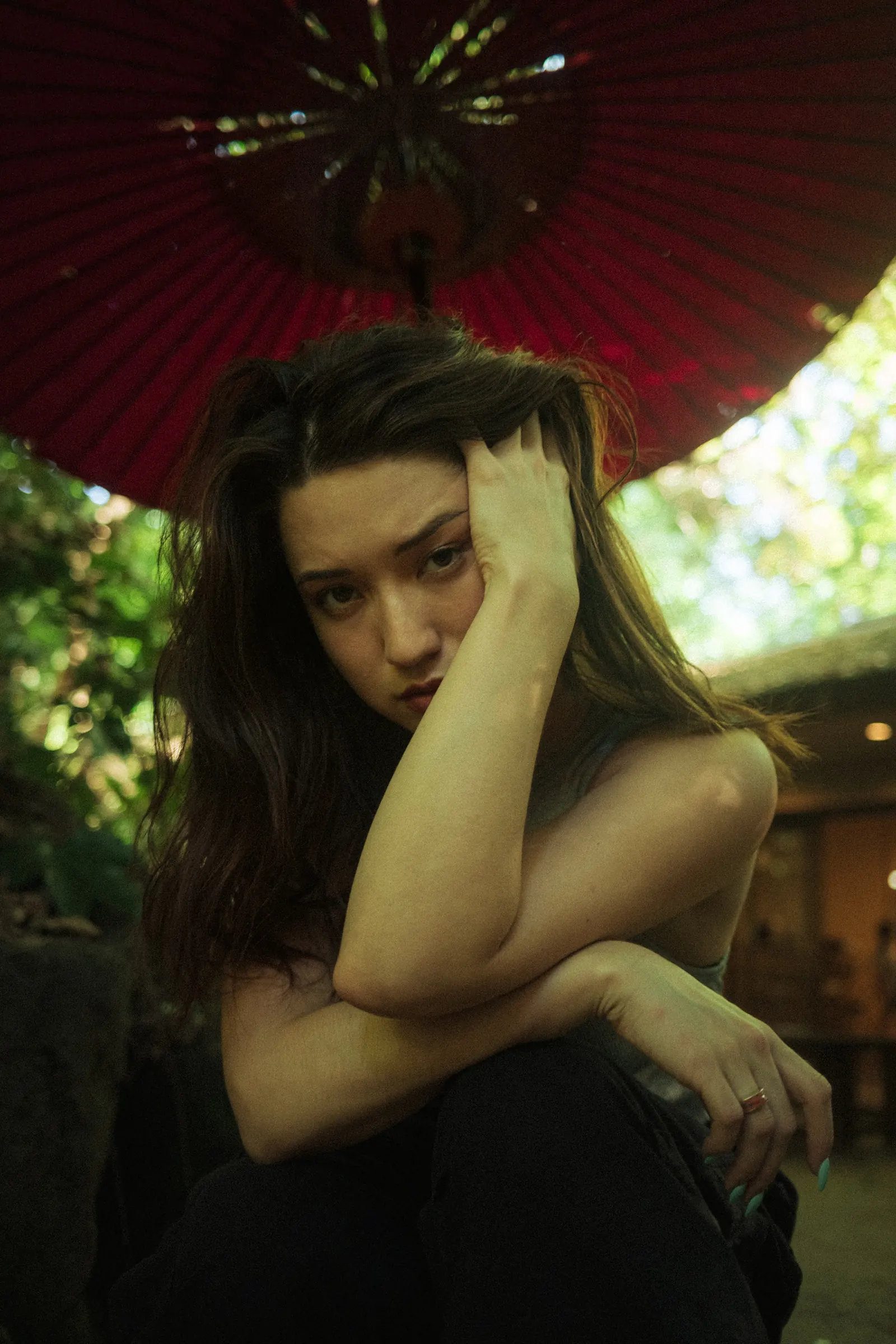 A woman with a hand in her hair and her arm across her knee, looking into the camera under a red paper parasol.