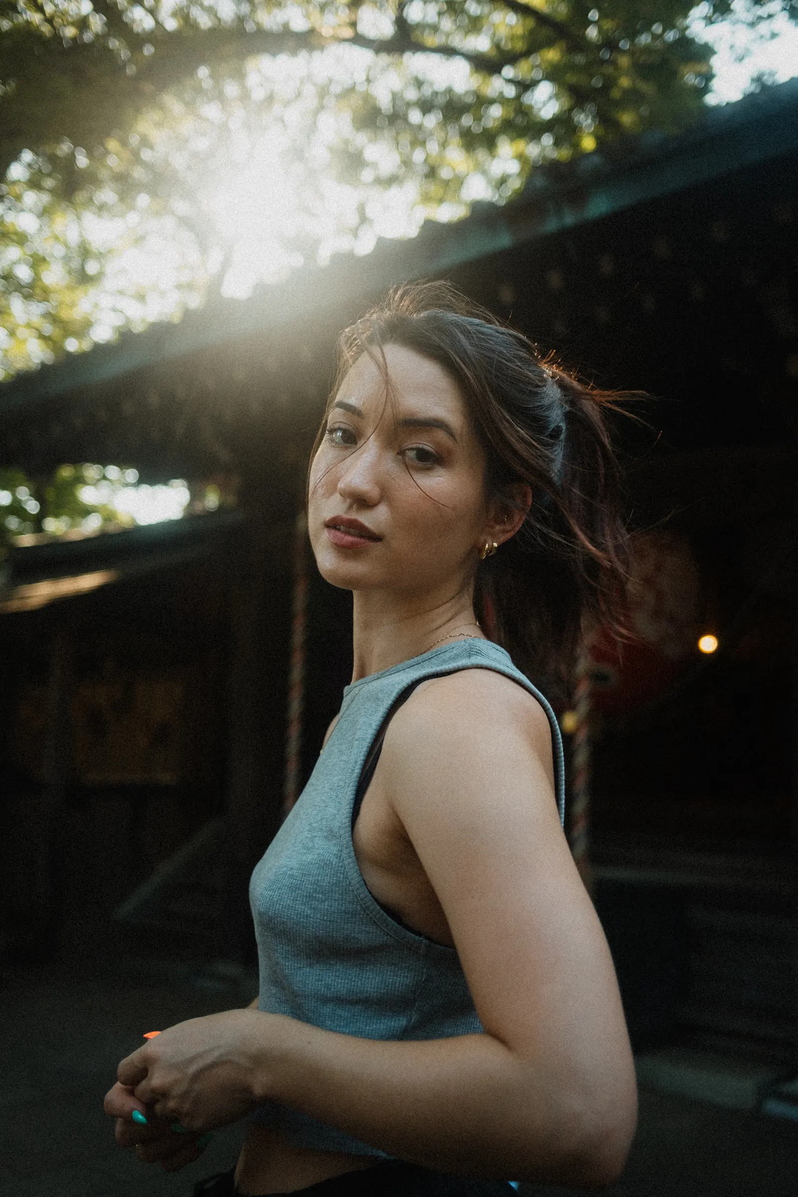 A woman looking back over her shoulder at the camera, hands clasped low, sunlight flaring through the trees above a dark shrine building.