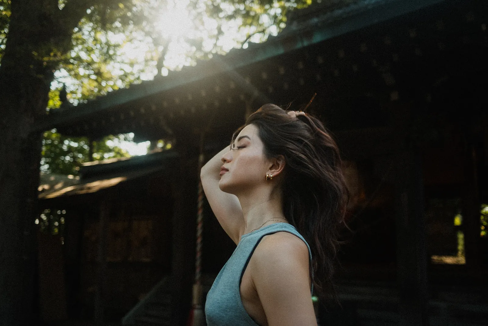 A woman in profile with her eyes closed and a hand in her windblown hair, sunlight breaking through the trees above a shrine.
