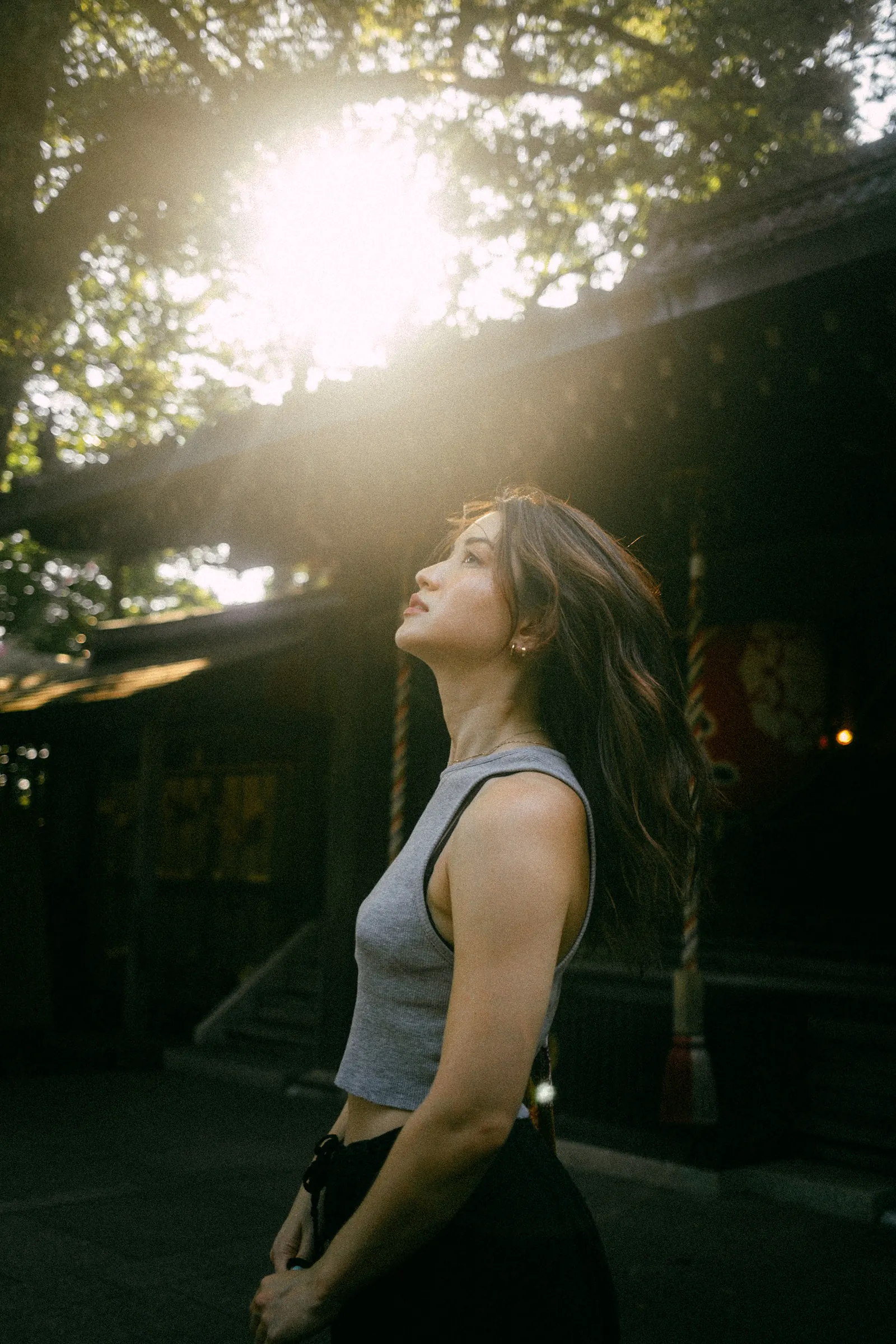 A woman in profile looking up into a burst of sunlight through the trees, a dark shrine building behind her.