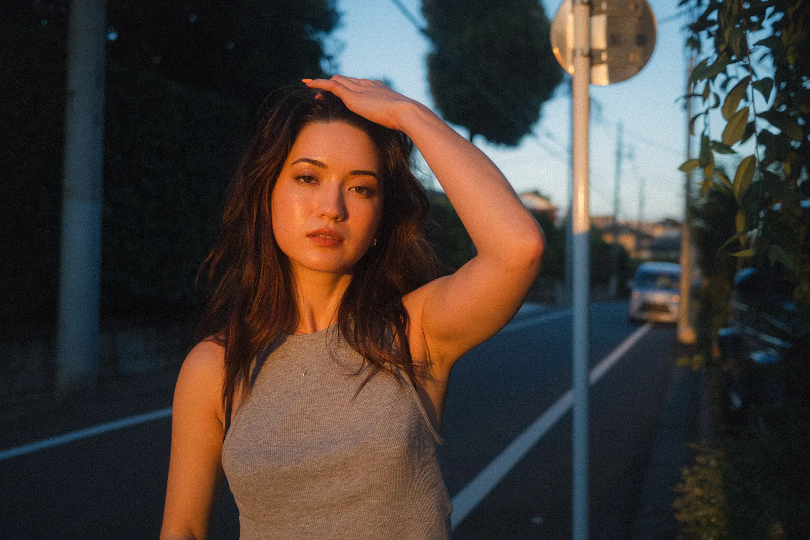 A woman with one hand on her head looking into the camera in warm evening light, a quiet road and a traffic mirror behind her.