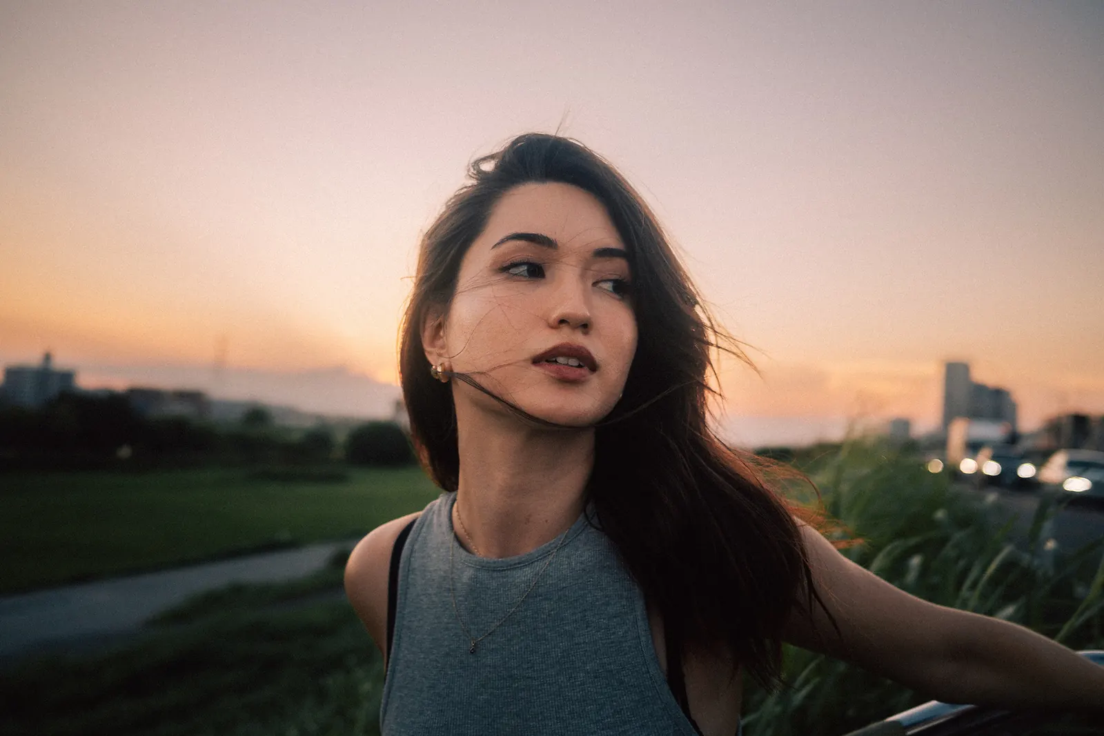A woman glancing away with wind in her hair at sunset, a grassy riverbank and soft car lights behind her.