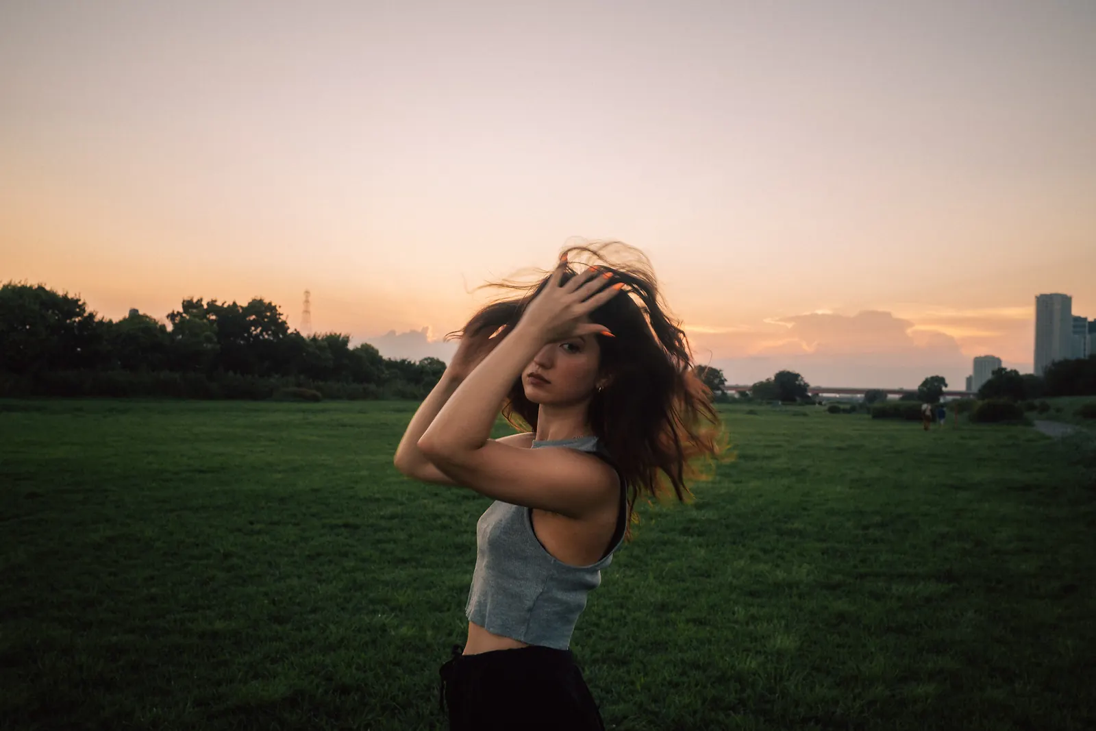 A woman with both hands in her tousled, windswept hair in an open field at dusk, city towers on the horizon.