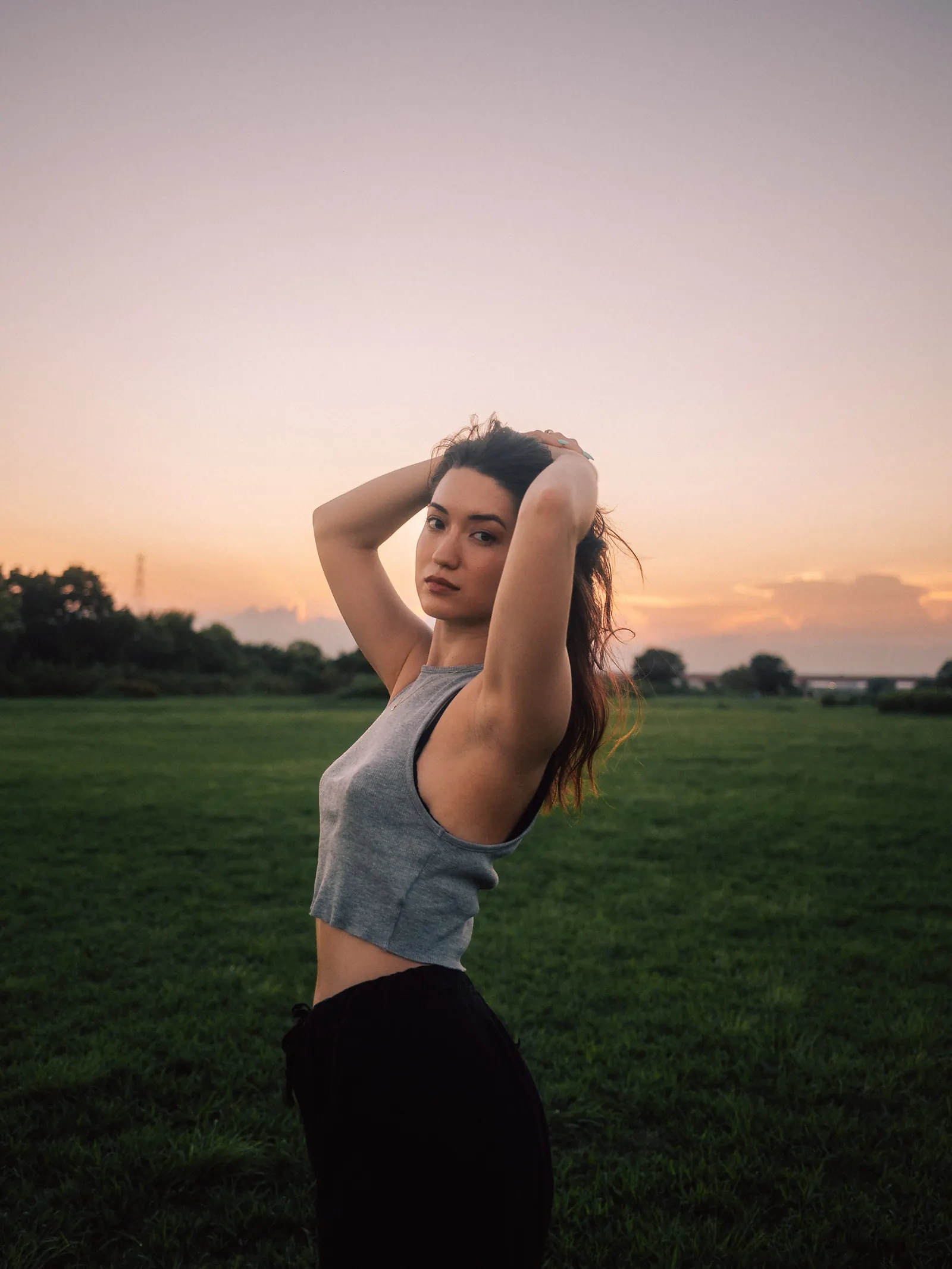 A woman looking over her shoulder at the camera with her hands behind her head, standing in a field at sunset.